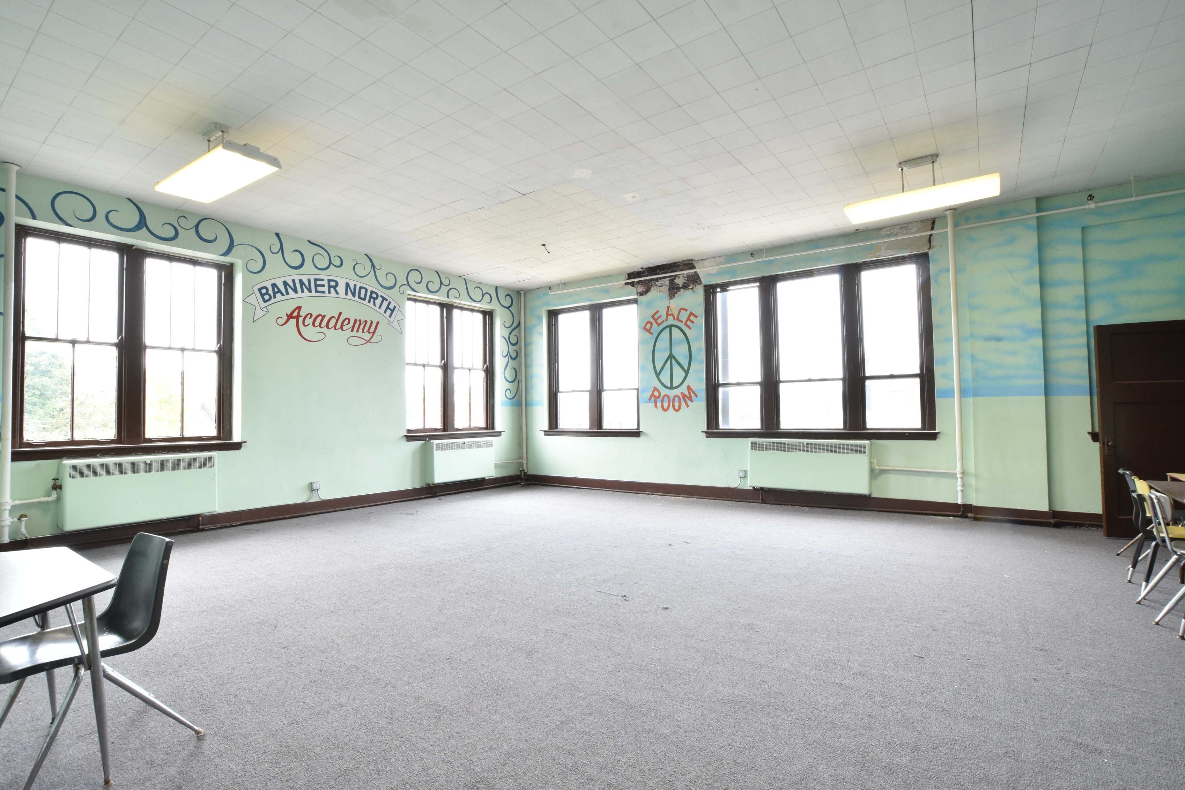 An empty classroom with large windows, light green walls featuring murals, and several tables and chairs arranged in the room.
