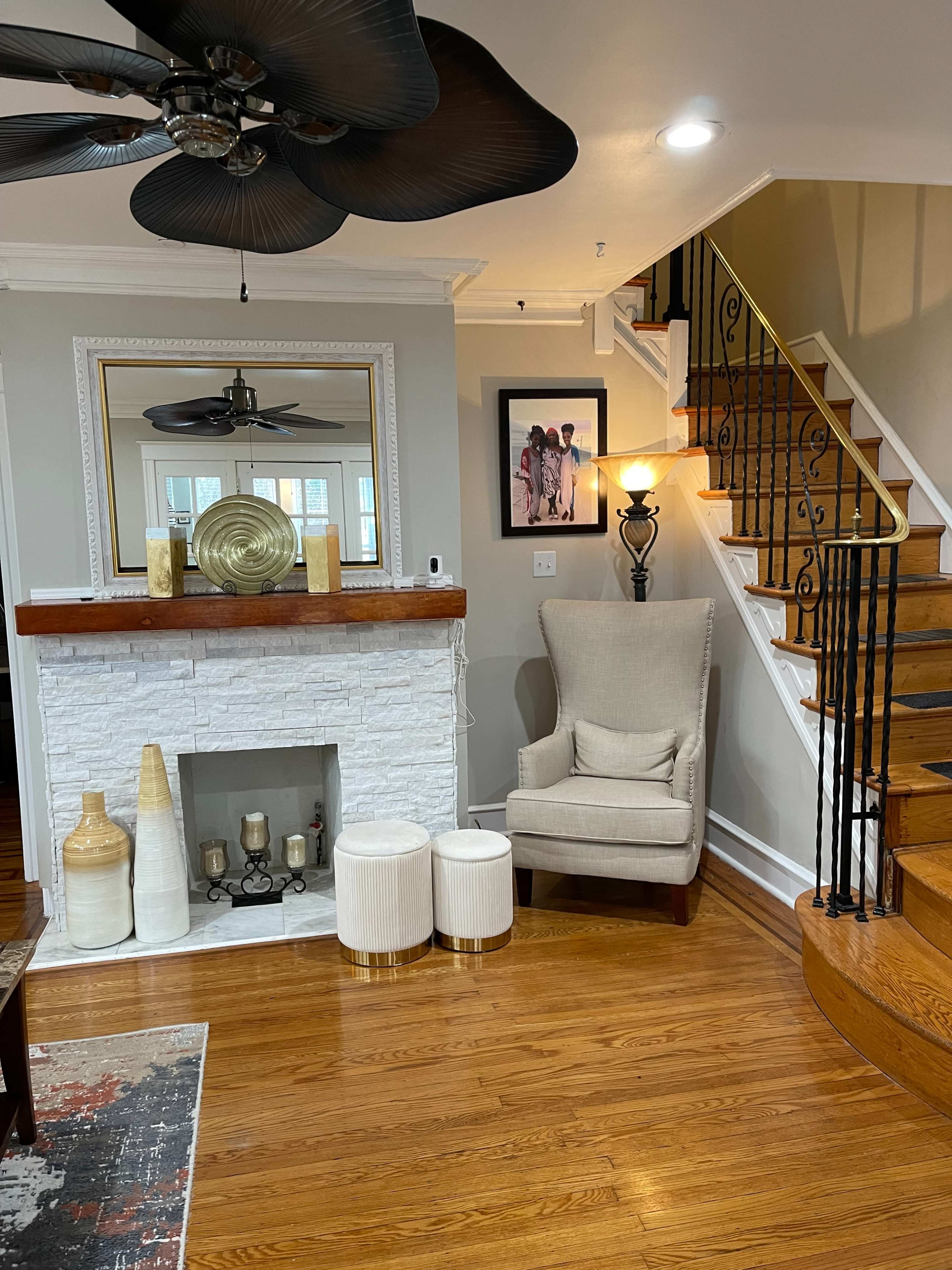 A living area featuring a stone fireplace with decorative items, a tall armchair, two small tables, a staircase, and artwork on the wall.