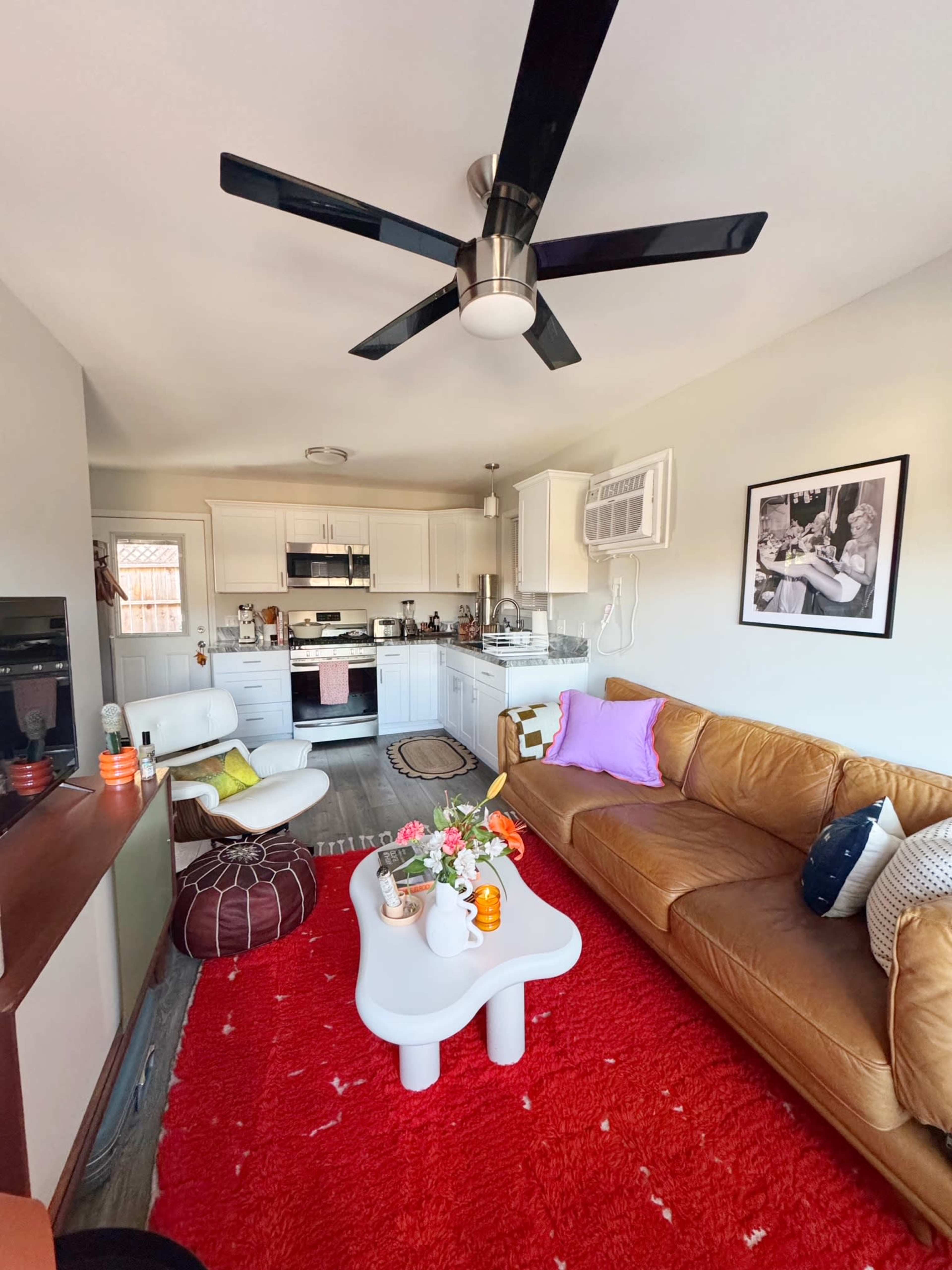 The image shows a living room with a brown leather sofa, colorful cushions, a white coffee table, and a red carpet, featuring a modern ceiling fan and kitchen area in the background.