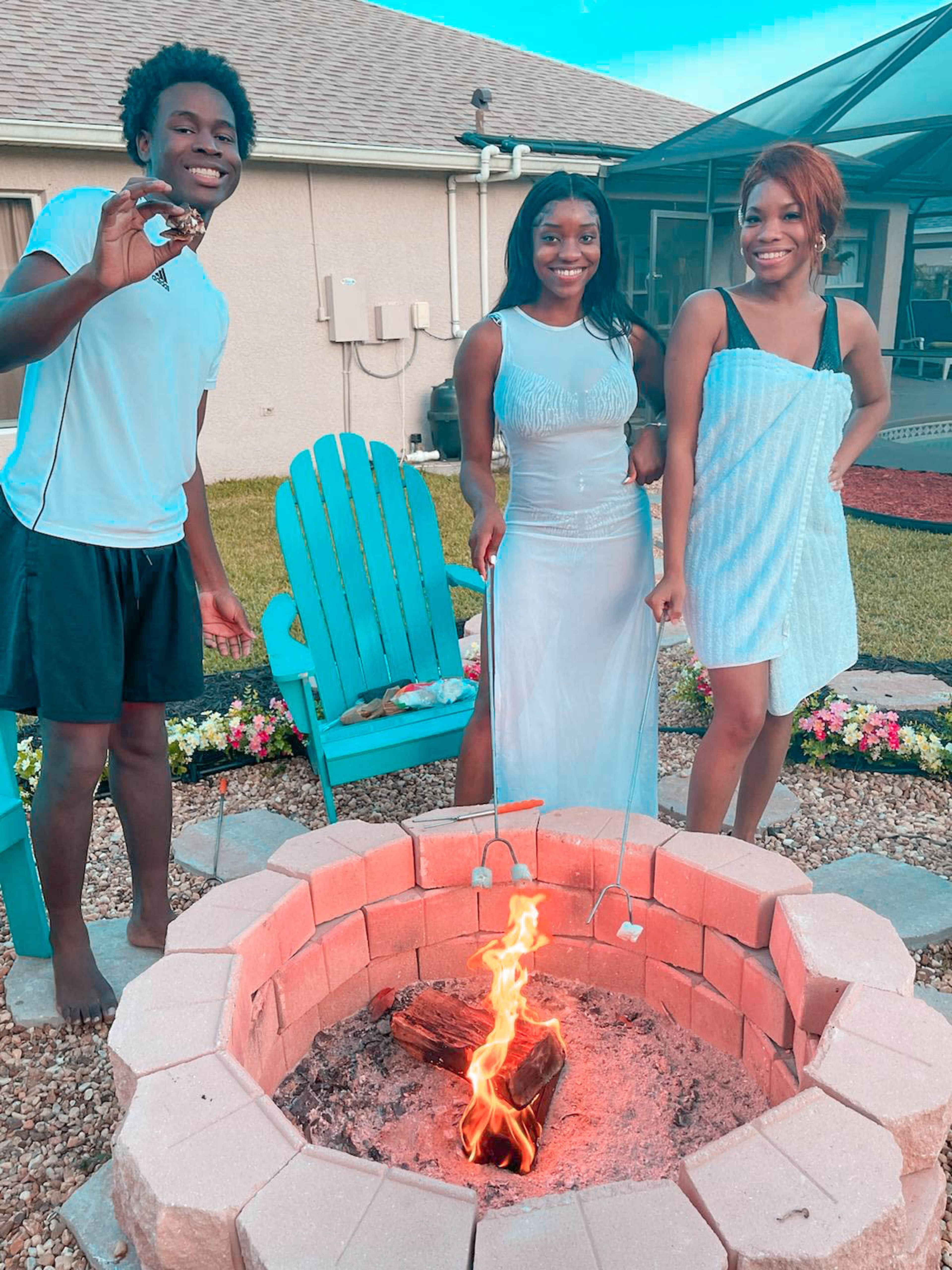 Three people stand around a fire pit, each holding a skewer, in a backyard setting with colorful chairs and flowers in the background.