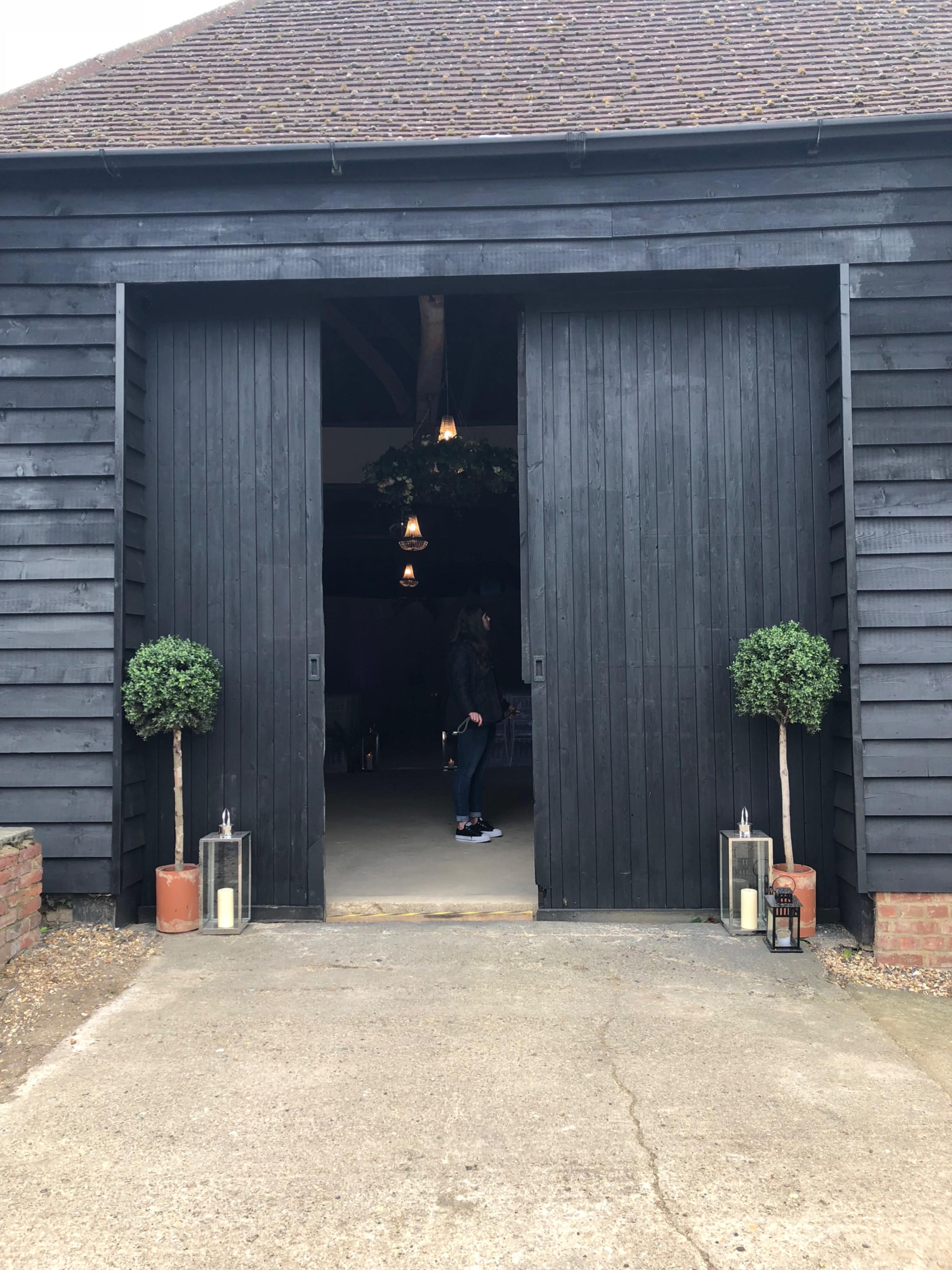 The image shows a large black wooden barn door, partially open, with a person inside and potted topiary plants on either side.