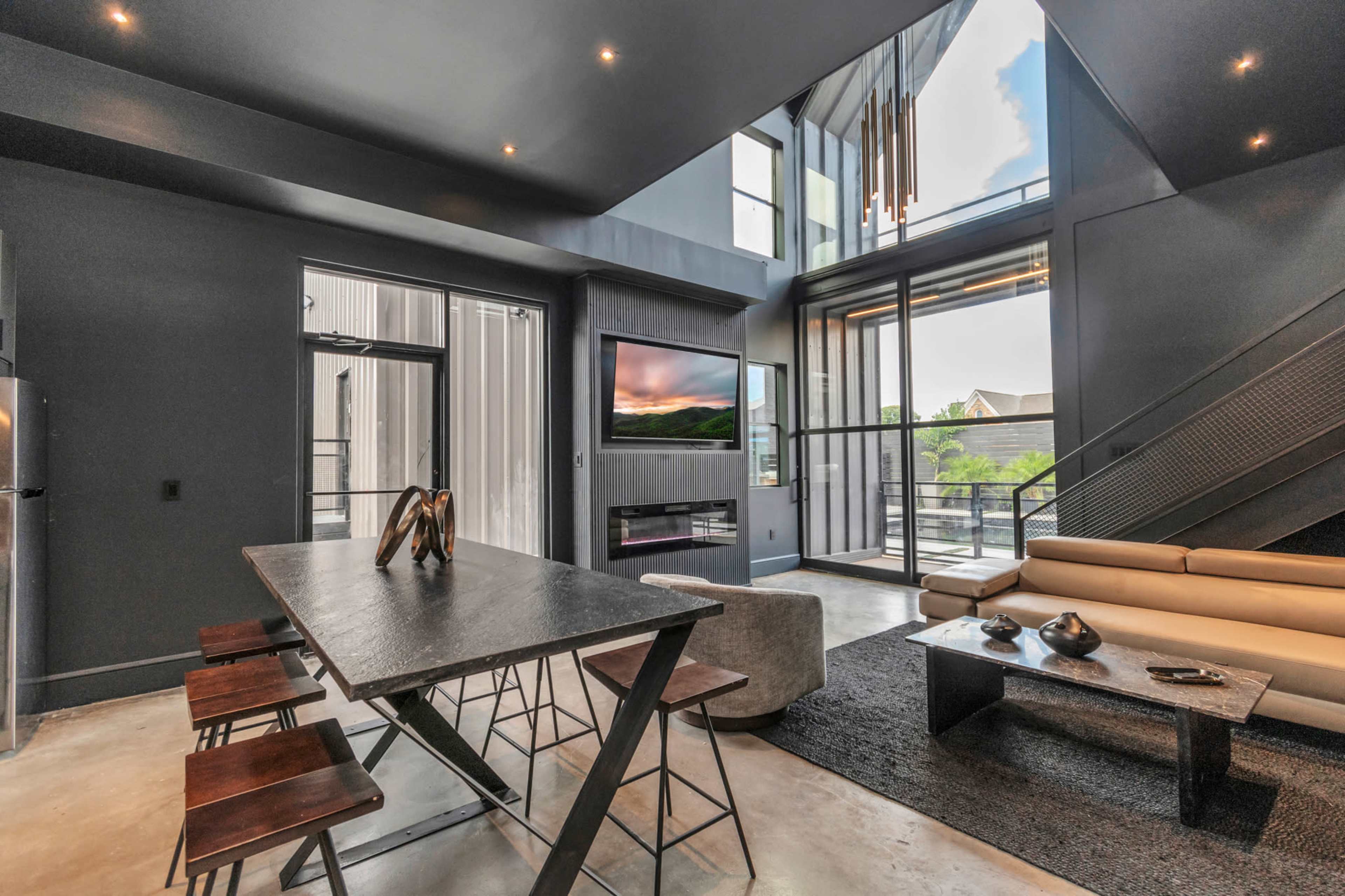 The image shows a modern living space with a high ceiling, large windows, a dining table, and a sectional sofa, overlooking an outdoor view.