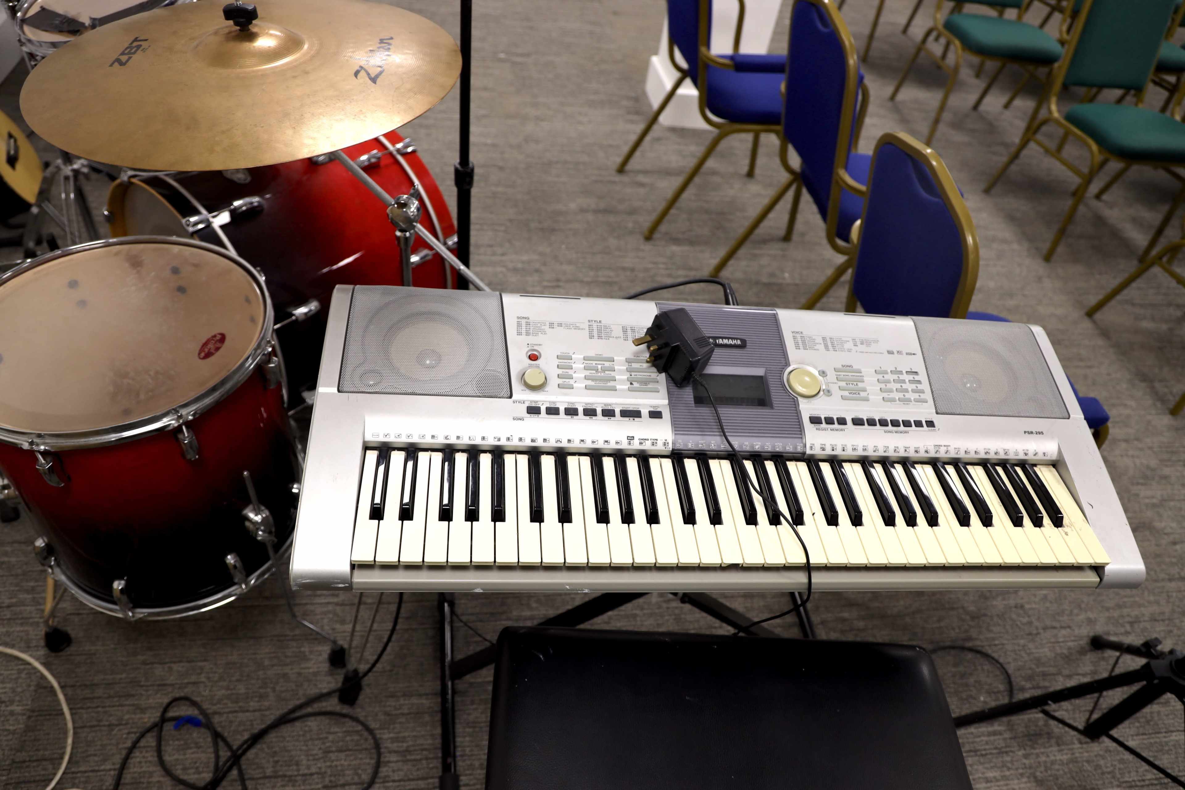 A digital keyboard is positioned next to a red drum set in a seating area with blue chairs.