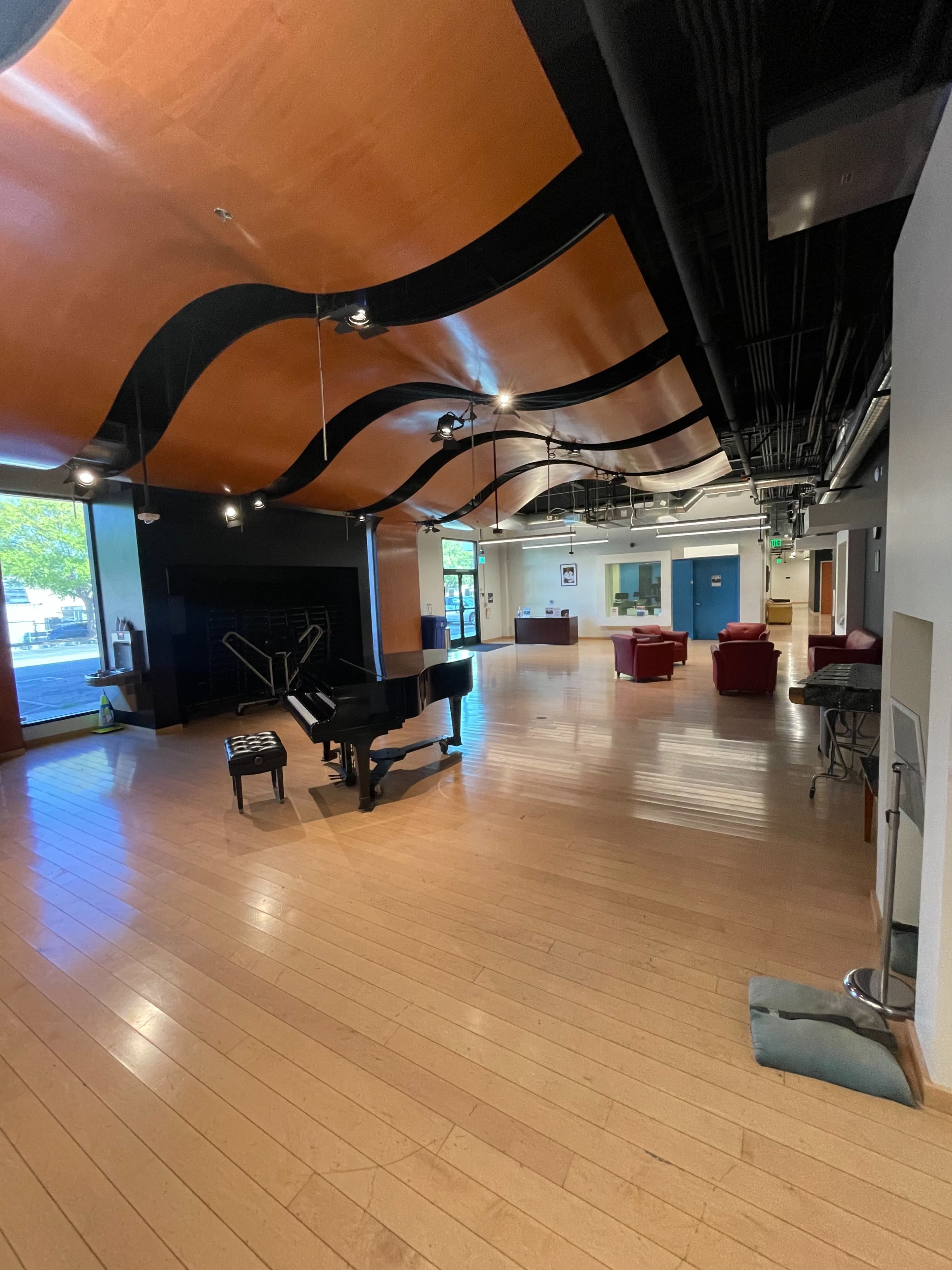 The image shows a spacious interior with wooden flooring, a grand piano positioned on the left, and seating arrangements on the right, all beneath a distinctive curved ceiling.
