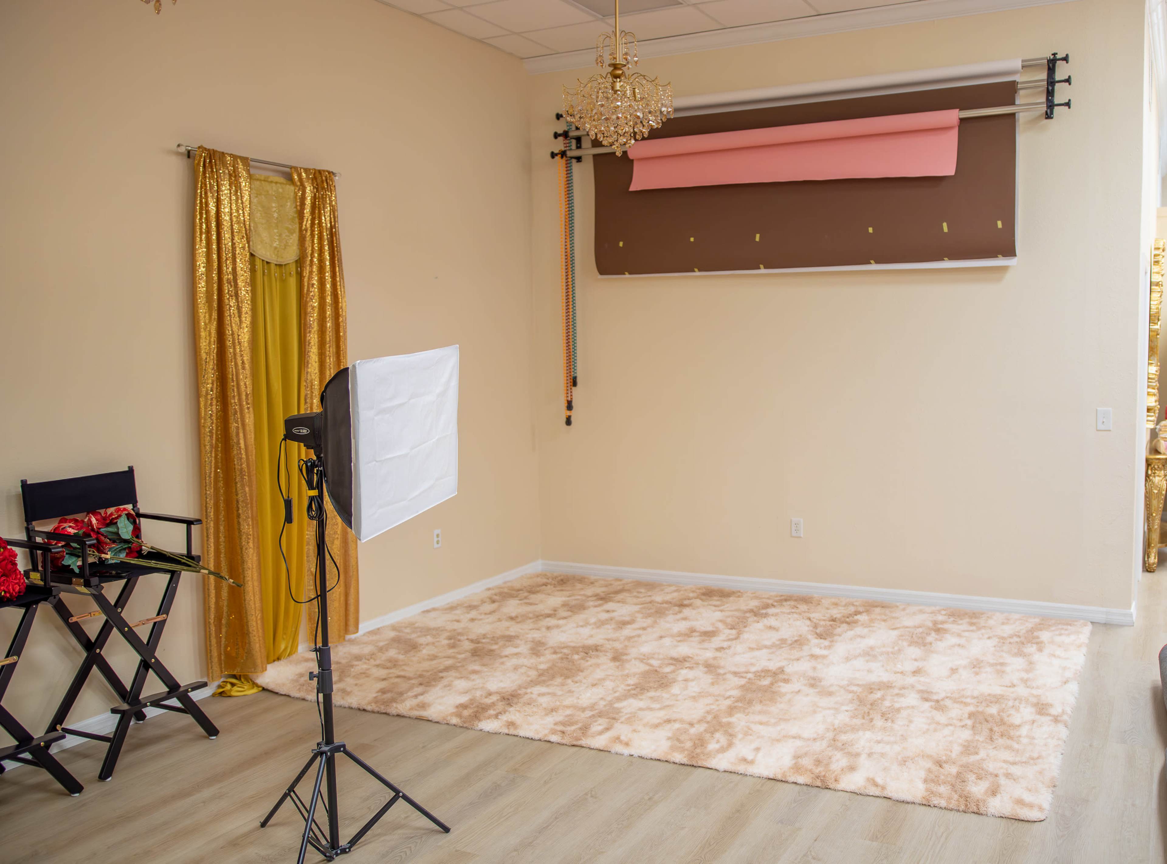 The image shows a light-colored room with a soft carpet, a photography backdrop setup on the wall, a standing light, and a director's chair near a gold curtain.