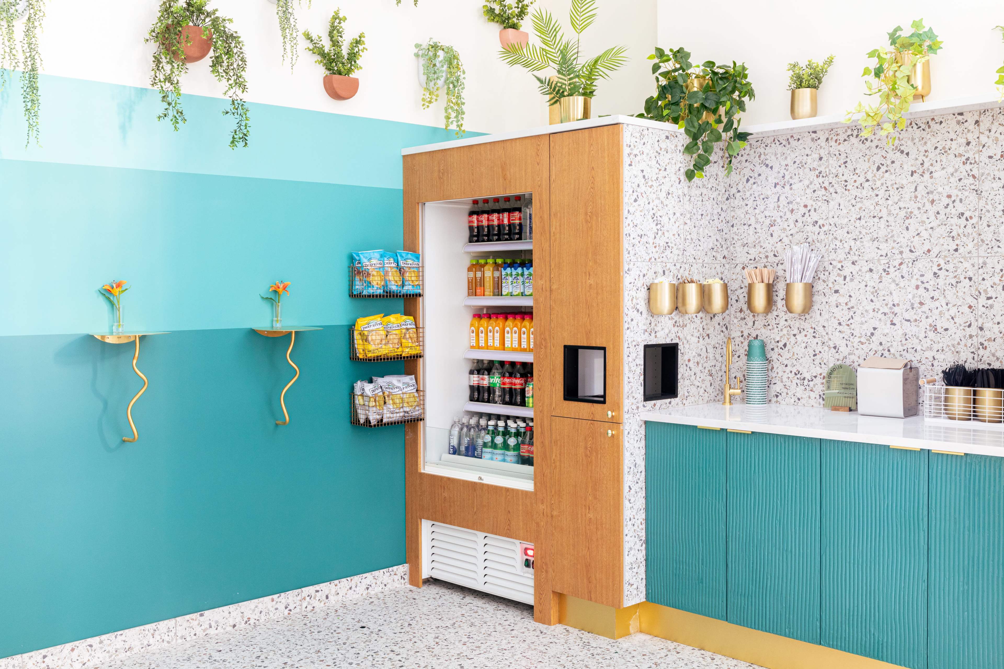 A modern snack and beverage station with a vending machine, shelving units, and a countertop adorned with plants and colorful items.