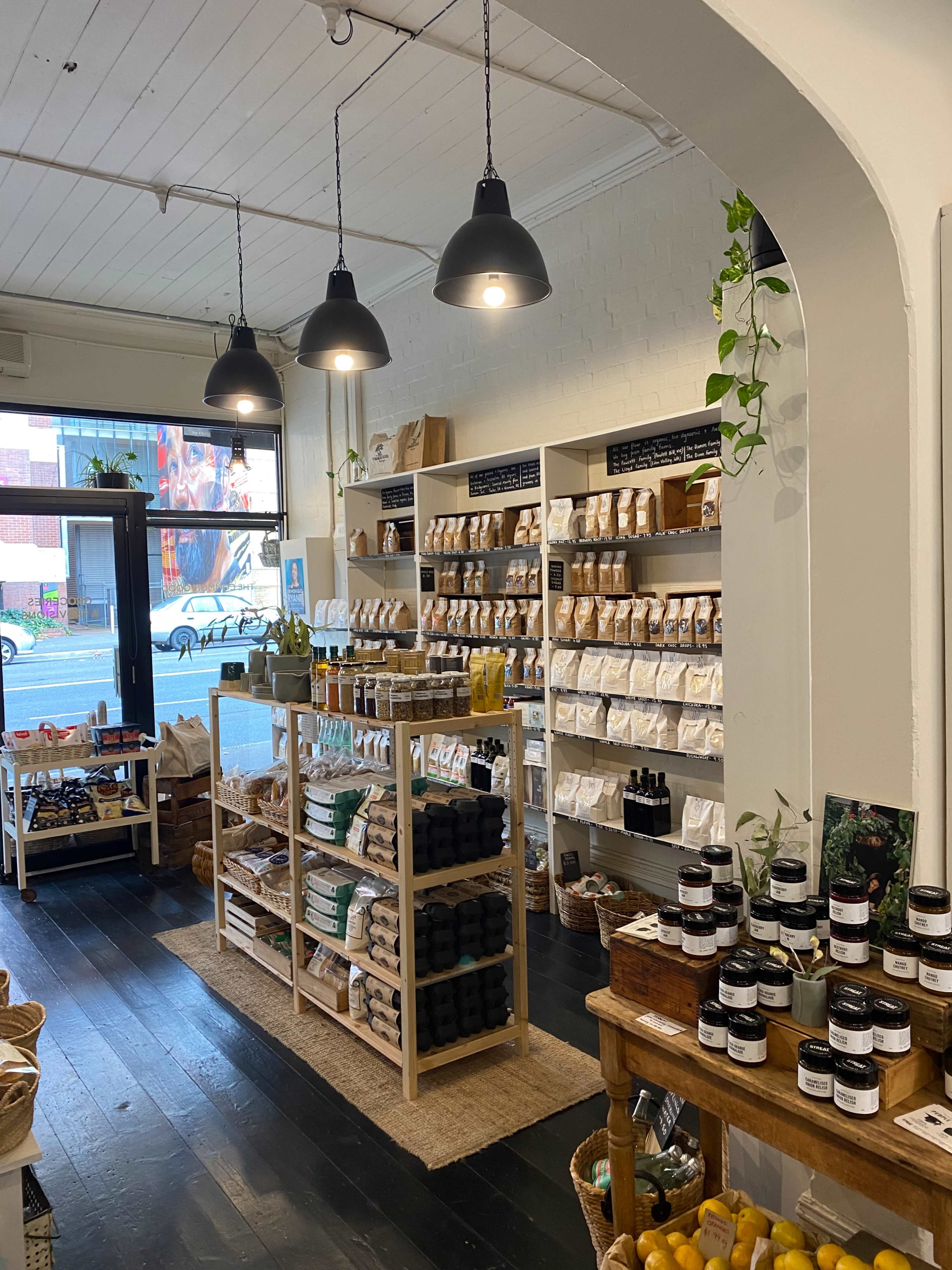 The image shows a well-organized retail space featuring shelves filled with various packaged goods, jars, and plants, illuminated by hanging black lights.