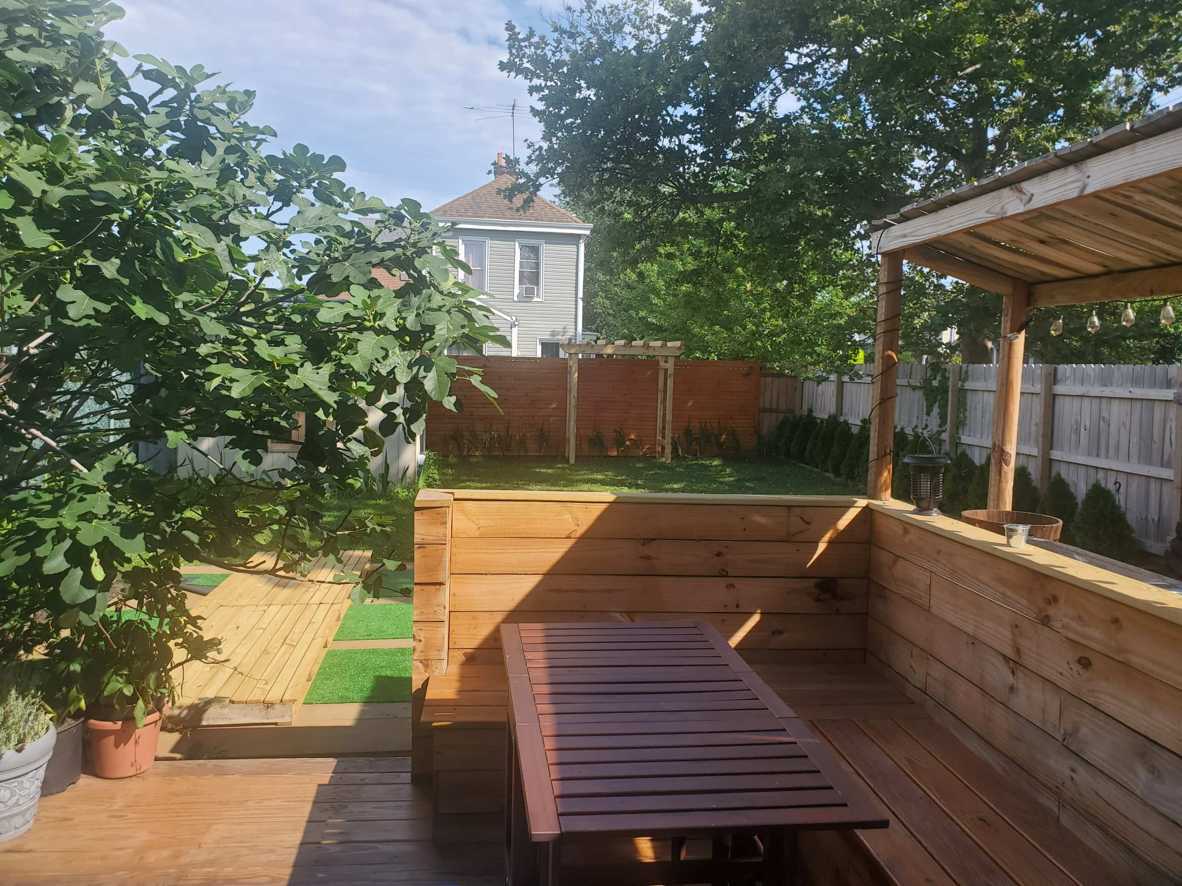 A wooden deck area with seating, surrounded by greenery and a landscaped yard, featuring a fence and a small structure in the background.
