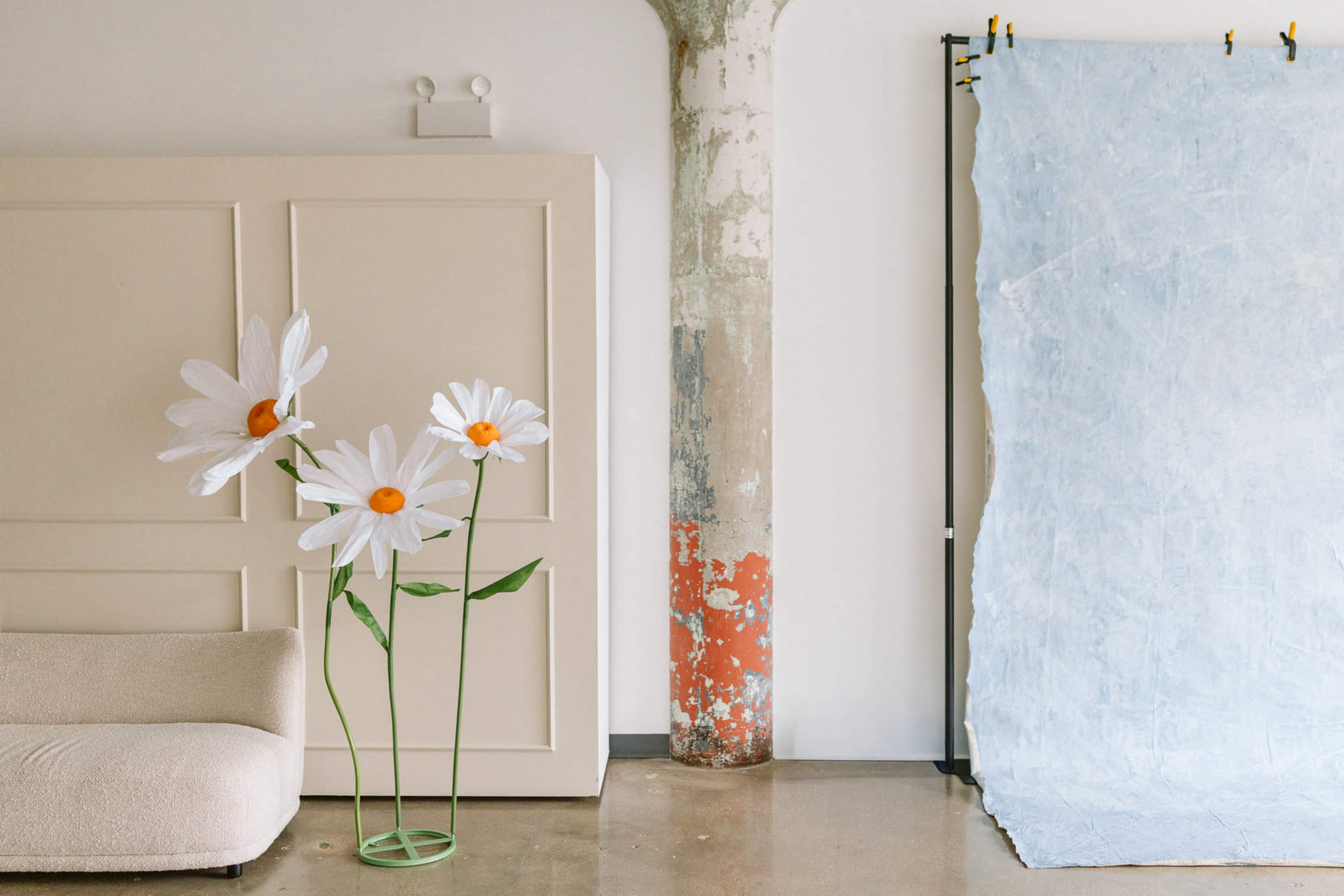 The image features large flower sculptures and a textured backdrop in a minimalist interior space.