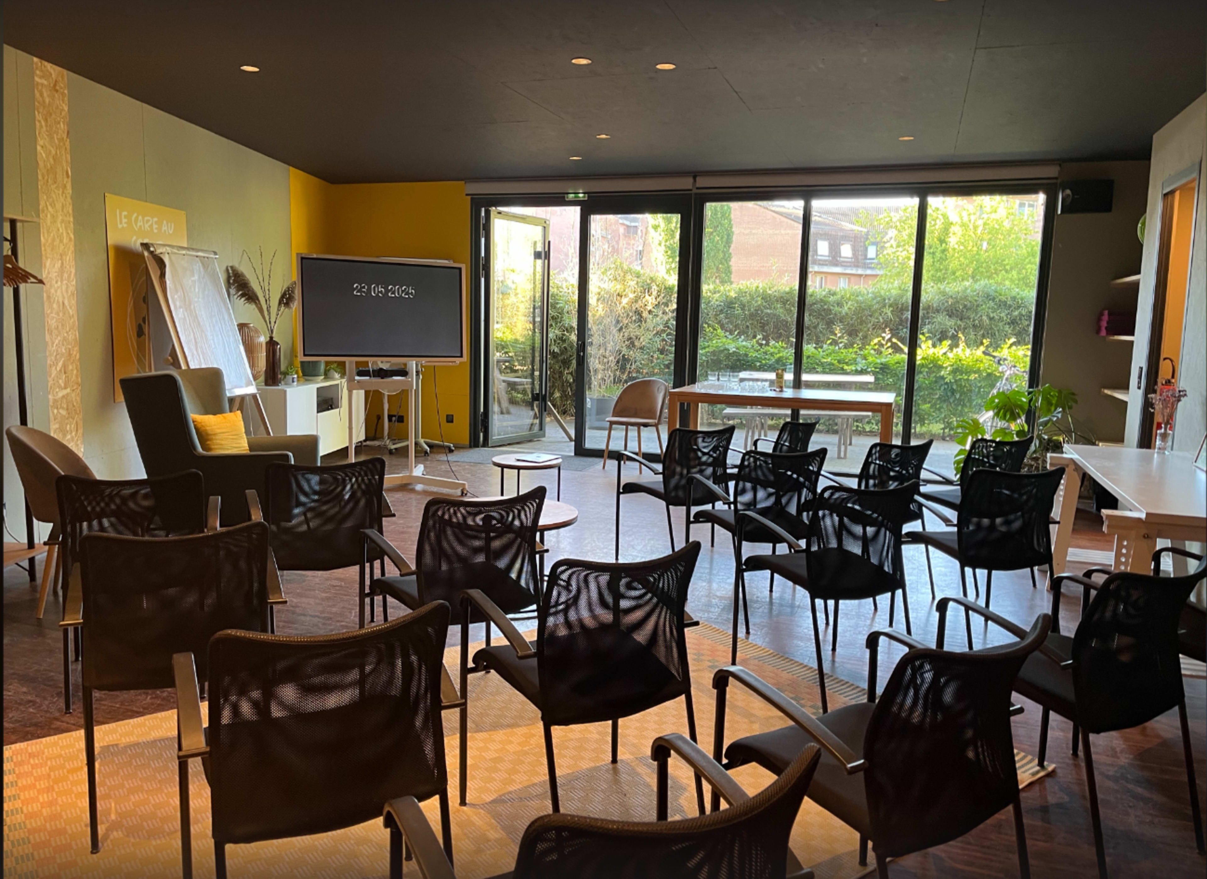 The image shows a modern conference room with rows of black chairs facing a screen, large windows revealing greenery outside, and a mix of yellow and white walls.