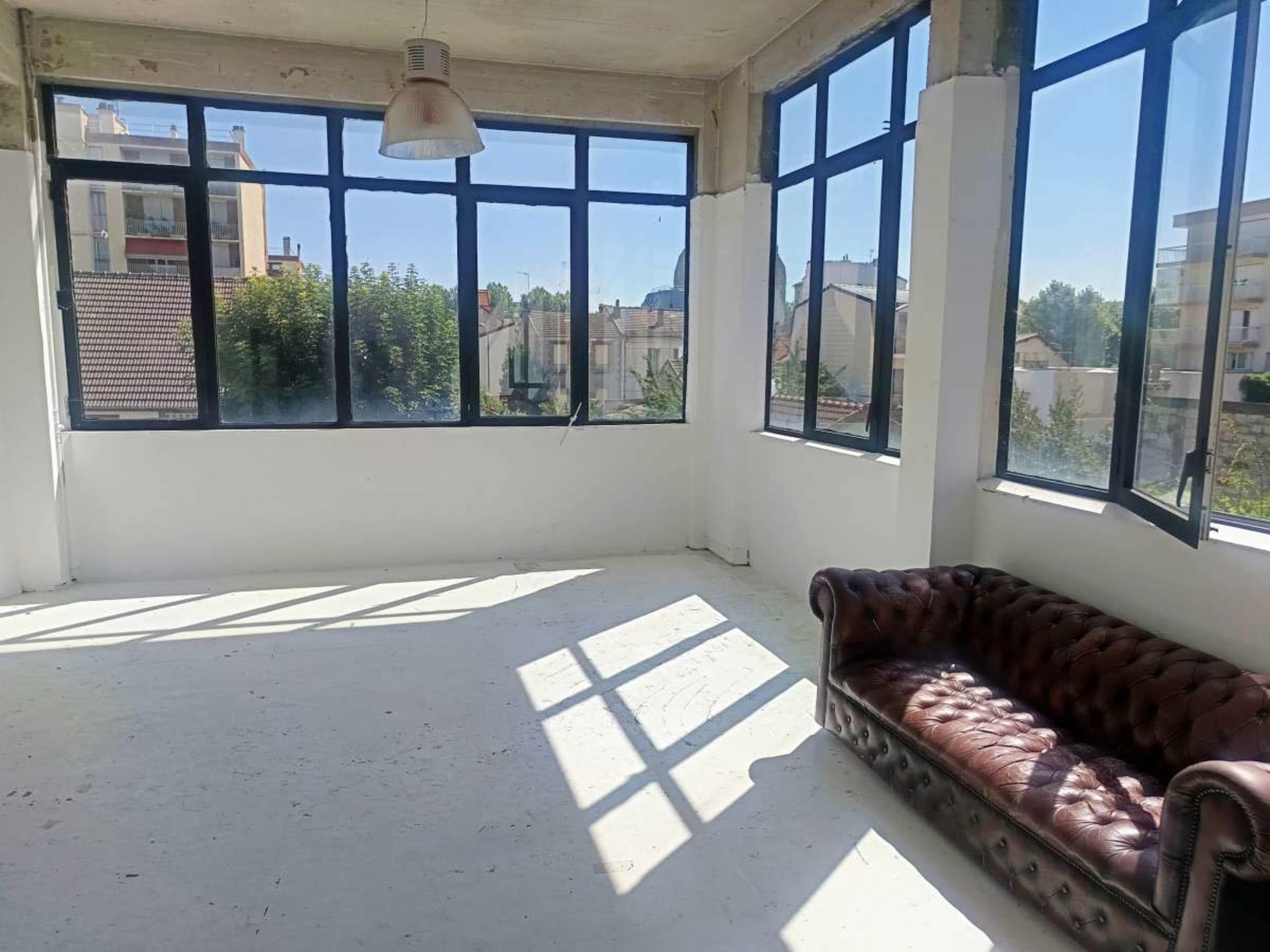 An empty room with large windows allowing sunlight to stream in, featuring a brown leather couch along the wall.