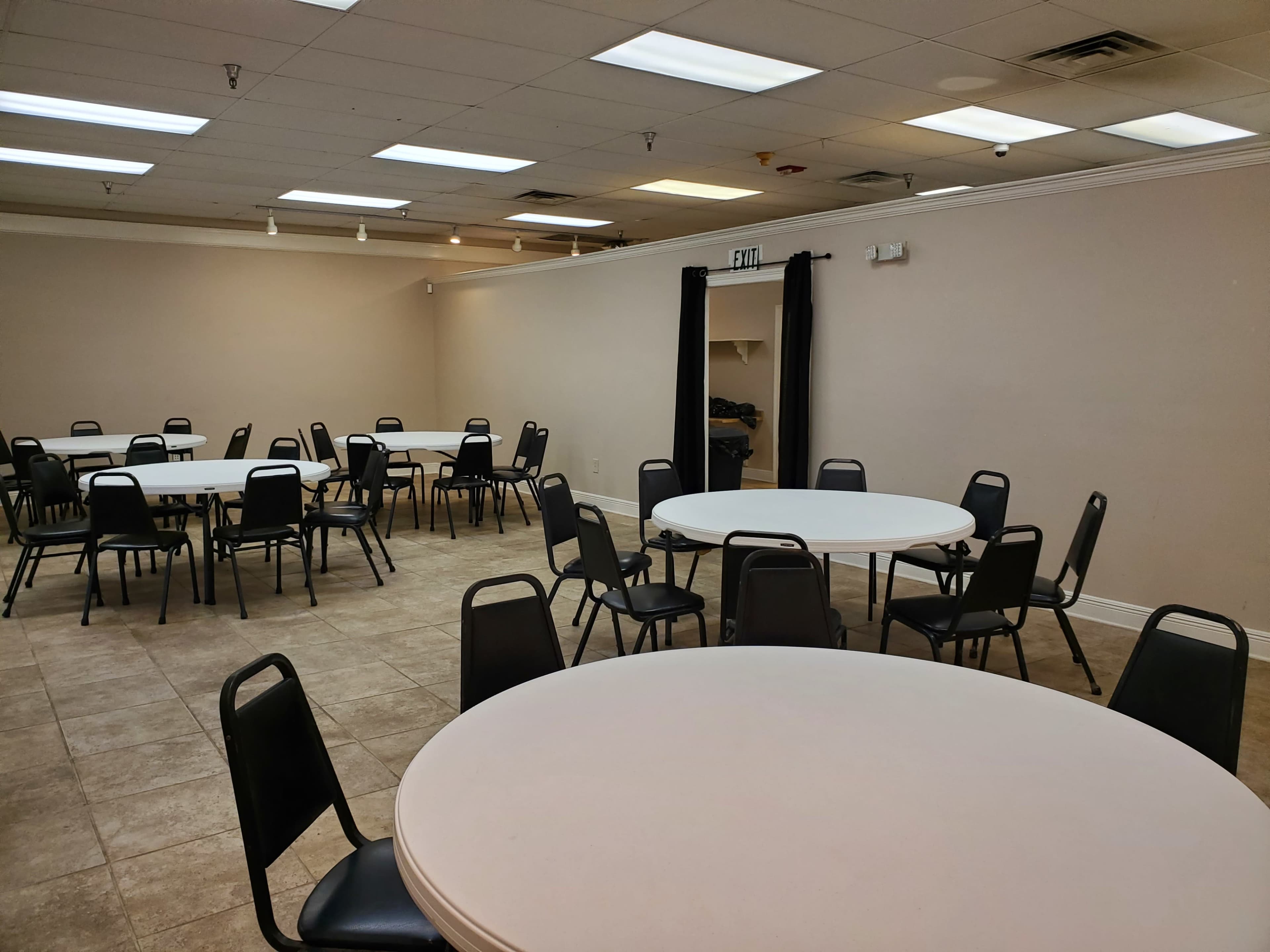 A large room with several round tables and chairs arranged throughout the space, featuring neutral-colored walls and tile flooring.