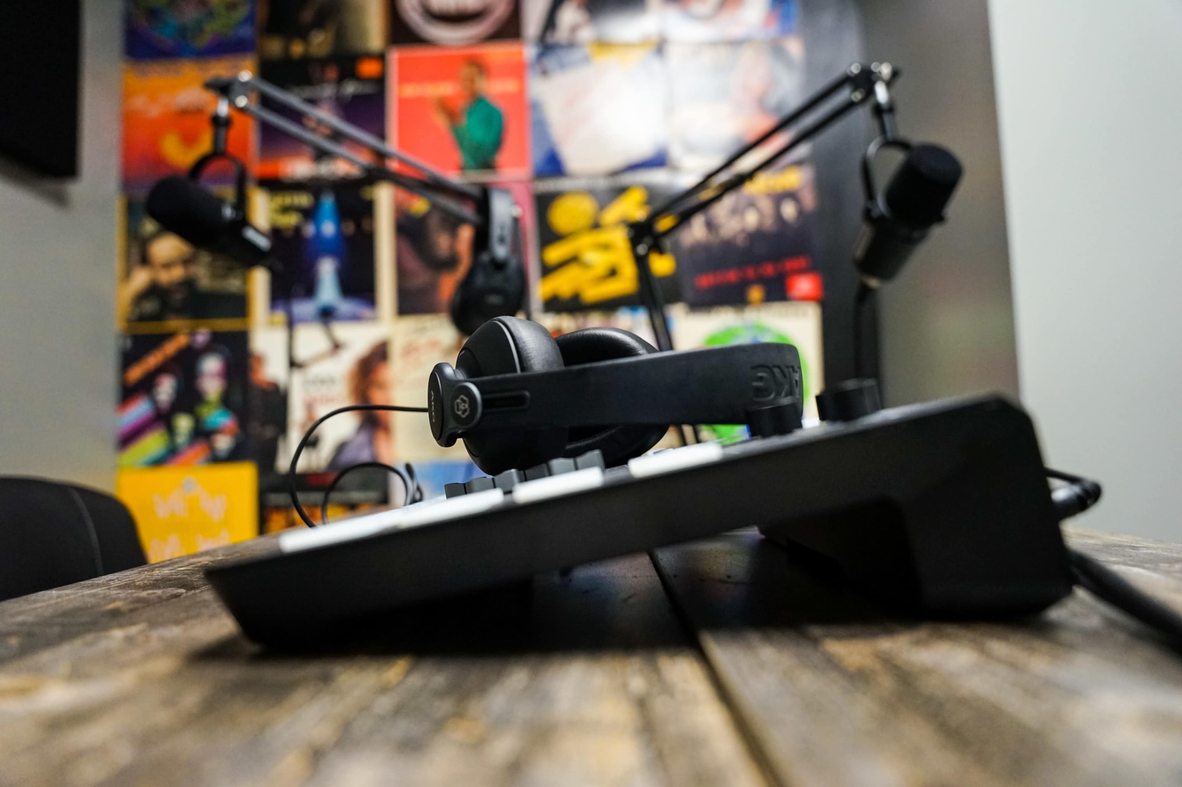 A pair of headphones rests on a tabletop in a room with colorful posters on the wall, and two microphones are mounted on adjustable arms.