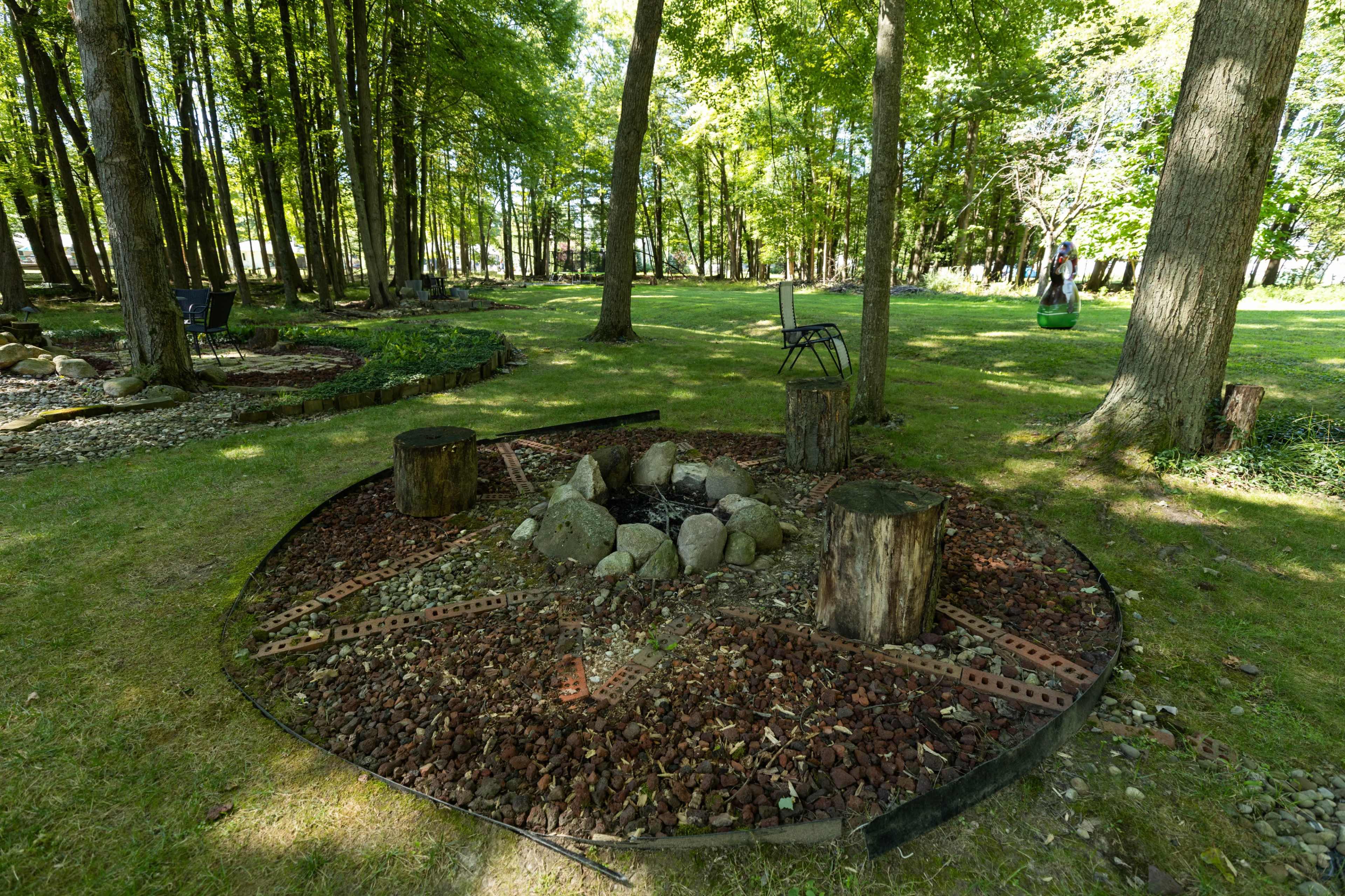 The image shows a circular fire pit area made of stones and logs, surrounded by a grassy lawn and trees.