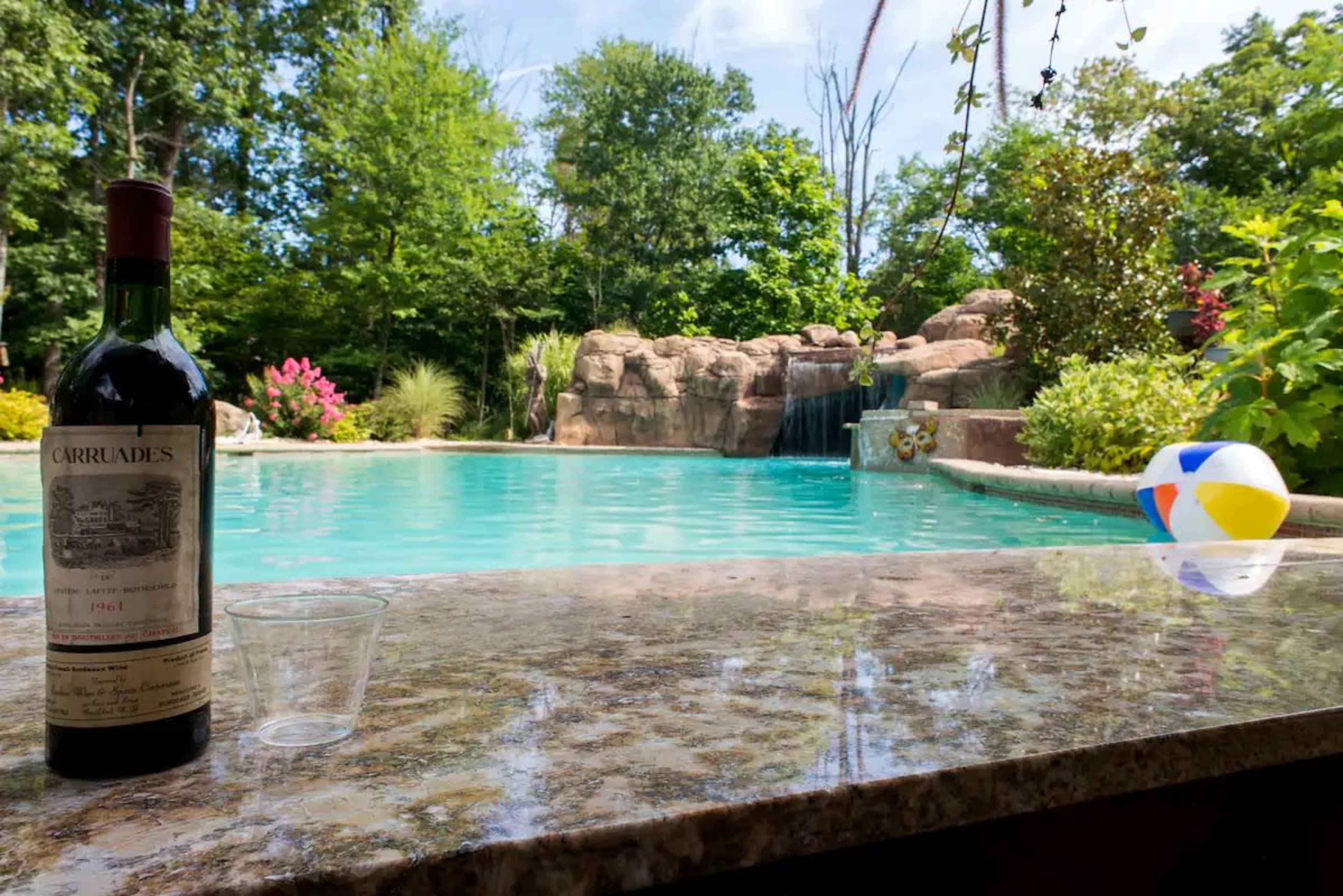 A bottle of wine and a clear plastic cup resting on a stone ledge beside a swimming pool surrounded by greenery and a waterfall.