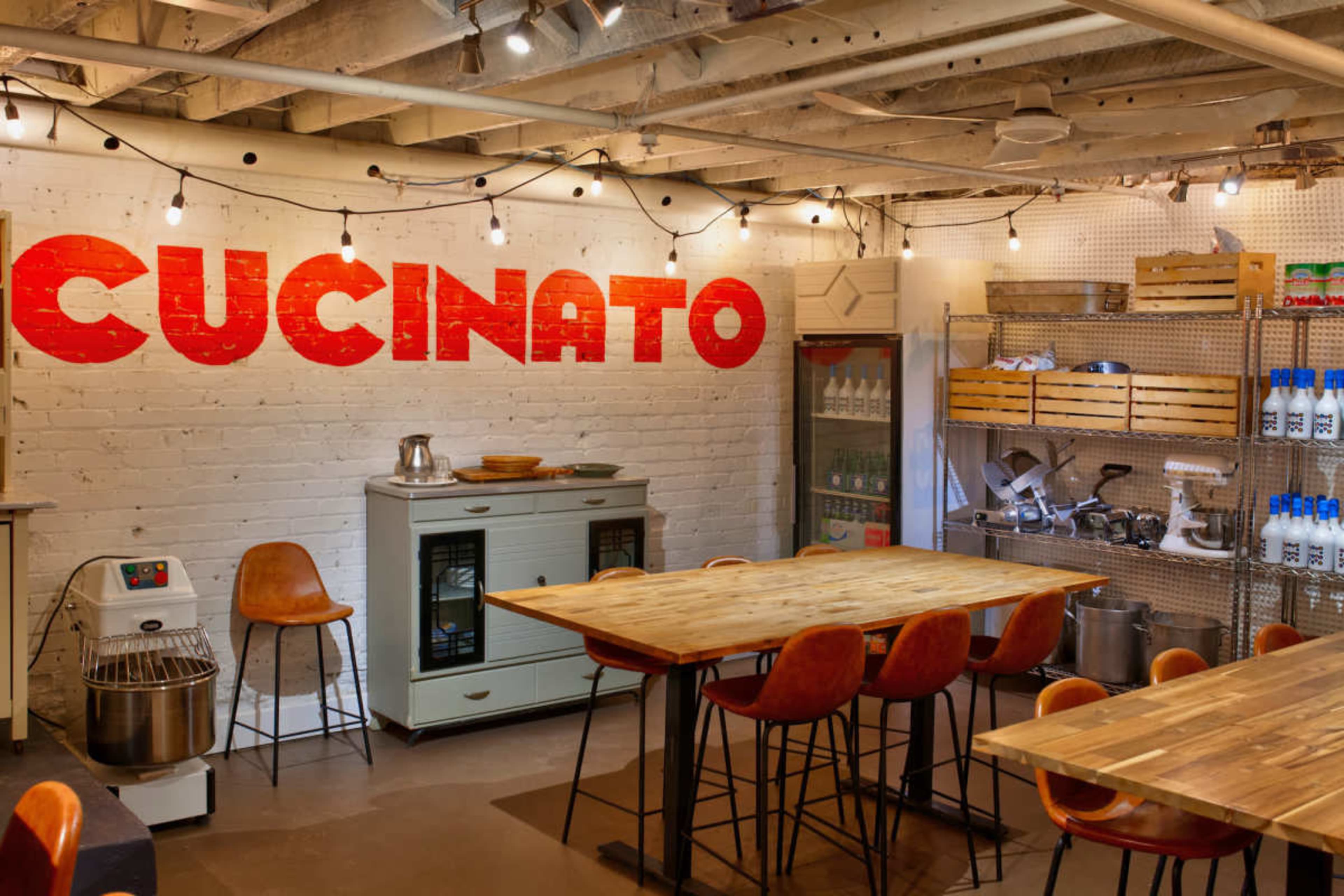 The image shows a brightly lit kitchen space with wooden tables, orange chairs, and a white brick wall featuring the word "CUCINATO" in large red letters.