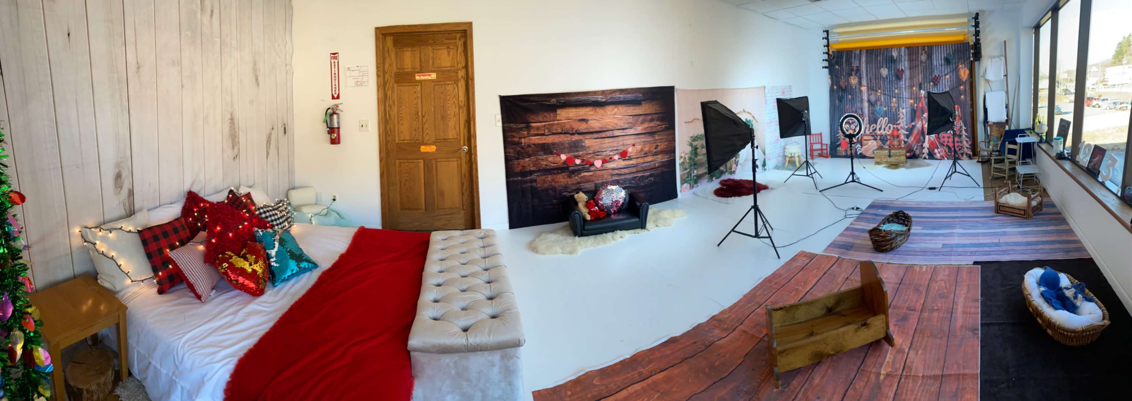 A spacious studio setup featuring a bed with decorative pillows, a Christmas tree, various backdrops, lighting equipment, and wooden props arranged on a white floor.