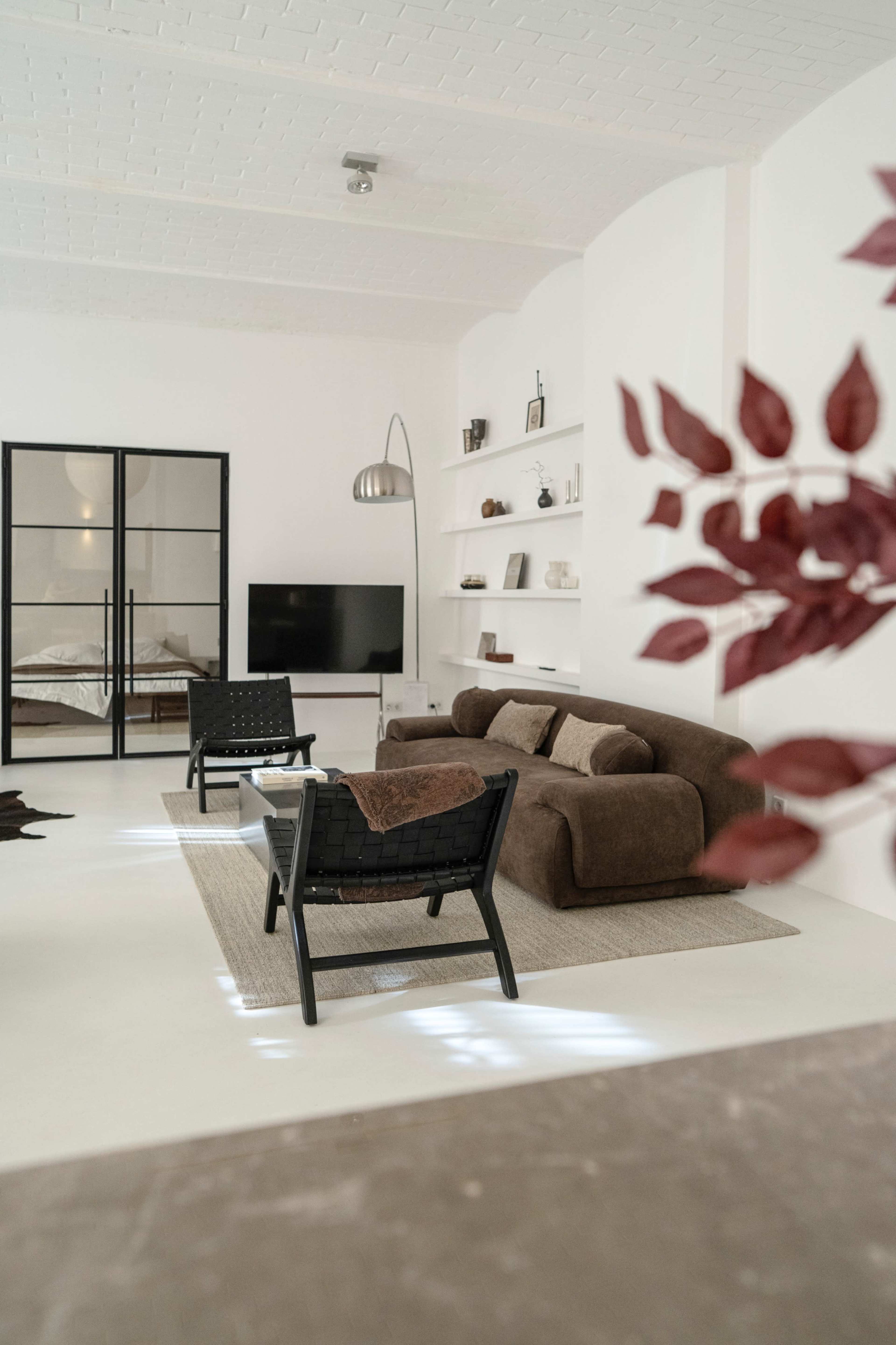 The image shows a modern, minimalist living room with a brown sofa, two black chairs, a television, and a shelving unit against a white wall.