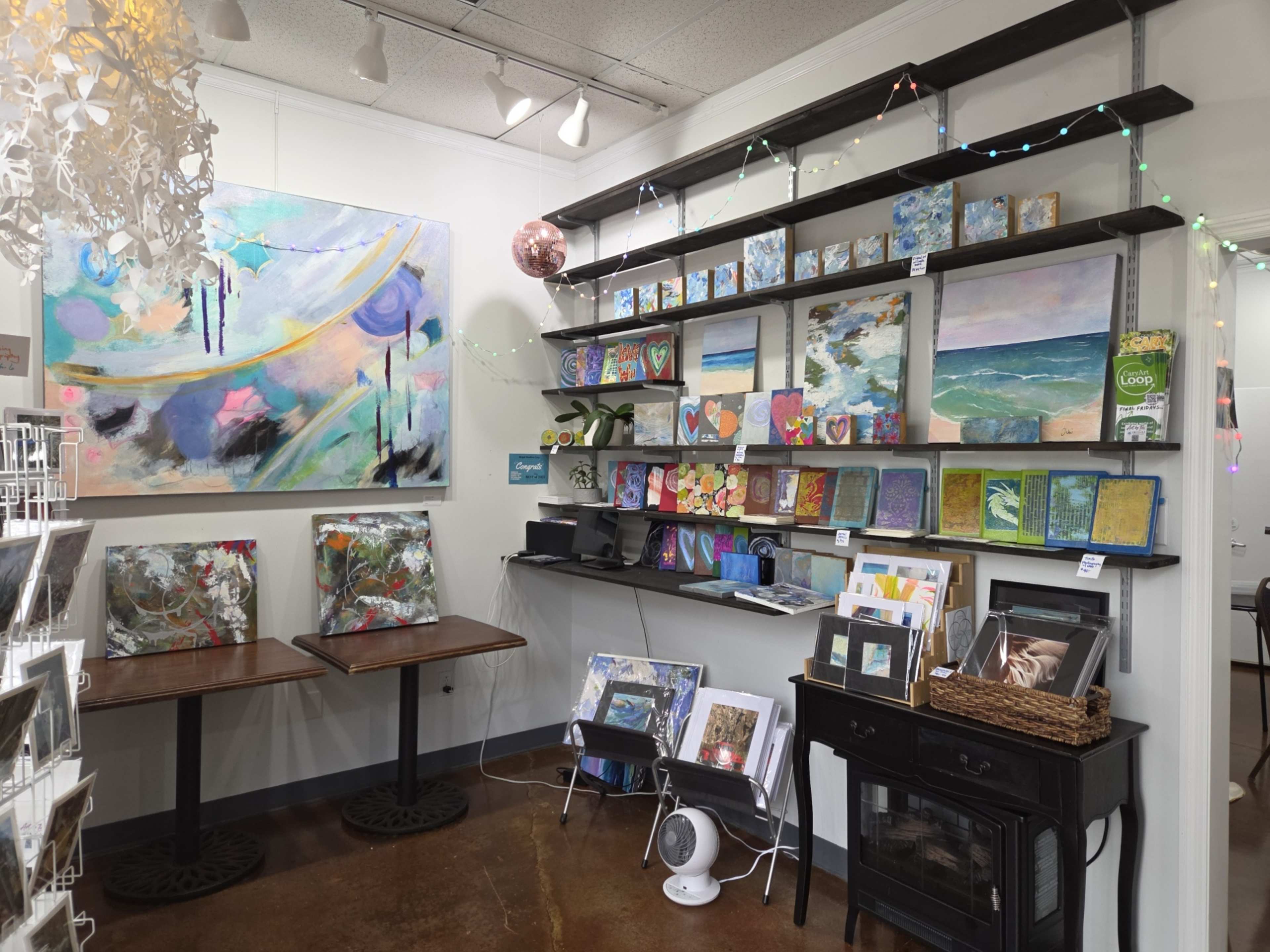 The image shows an art display with colorful paintings and artworks arranged on shelves and tables in a well-lit studio space.