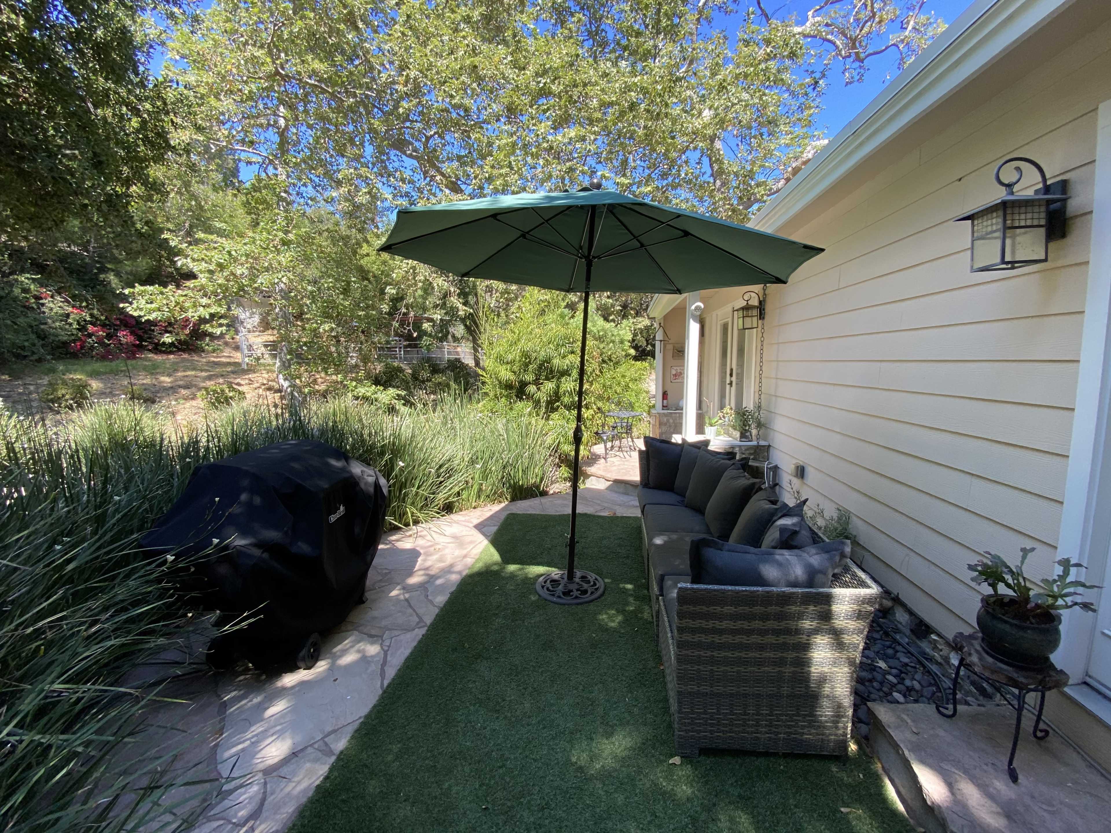 A patio area features a green umbrella over a seating arrangement and a grill beside a neatly landscaped garden.