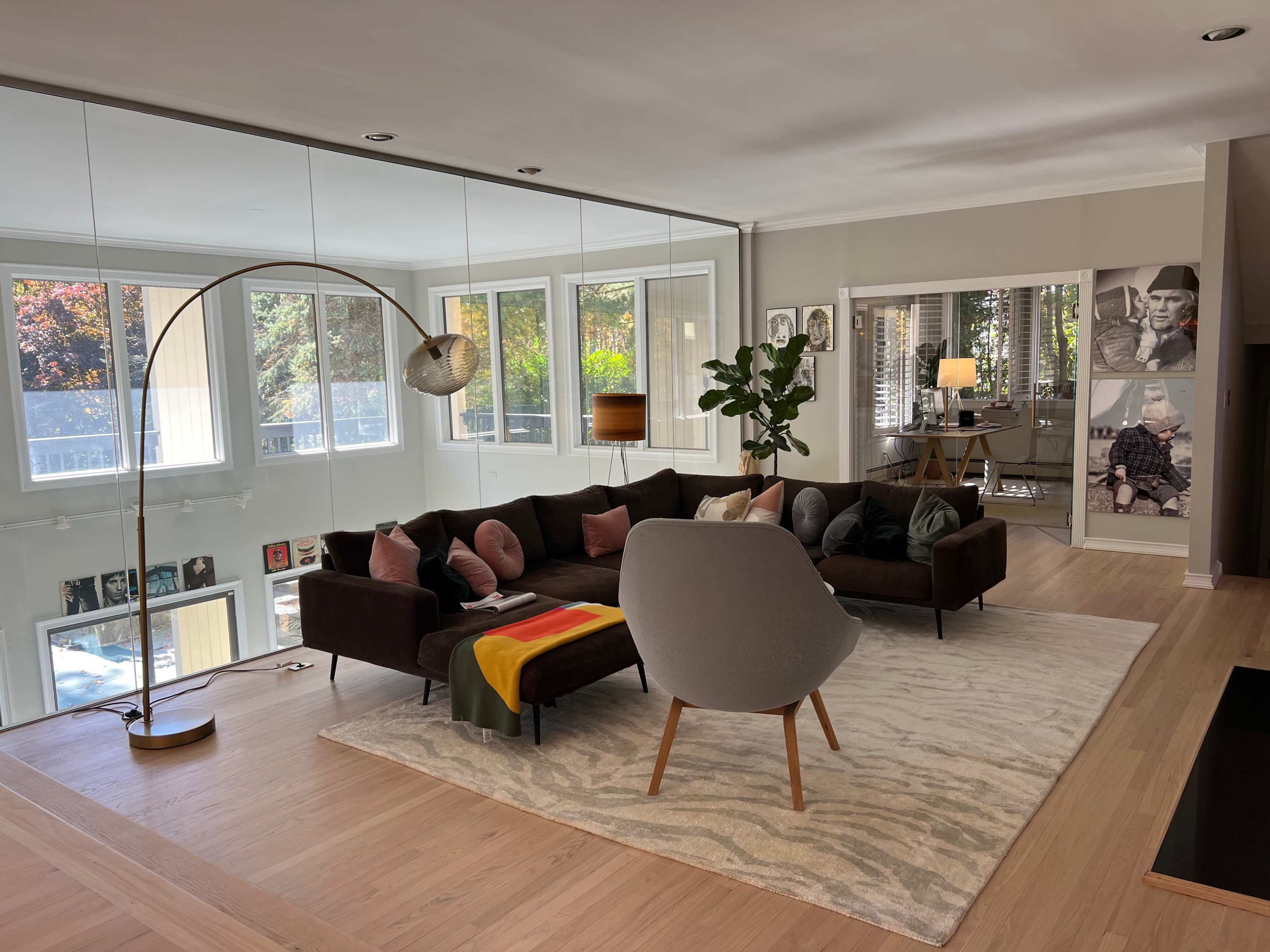 The image shows a modern living room featuring a large sectional sofa, a curved chair, a floor lamp, and large windows that provide natural light, with framed photographs on the walls.