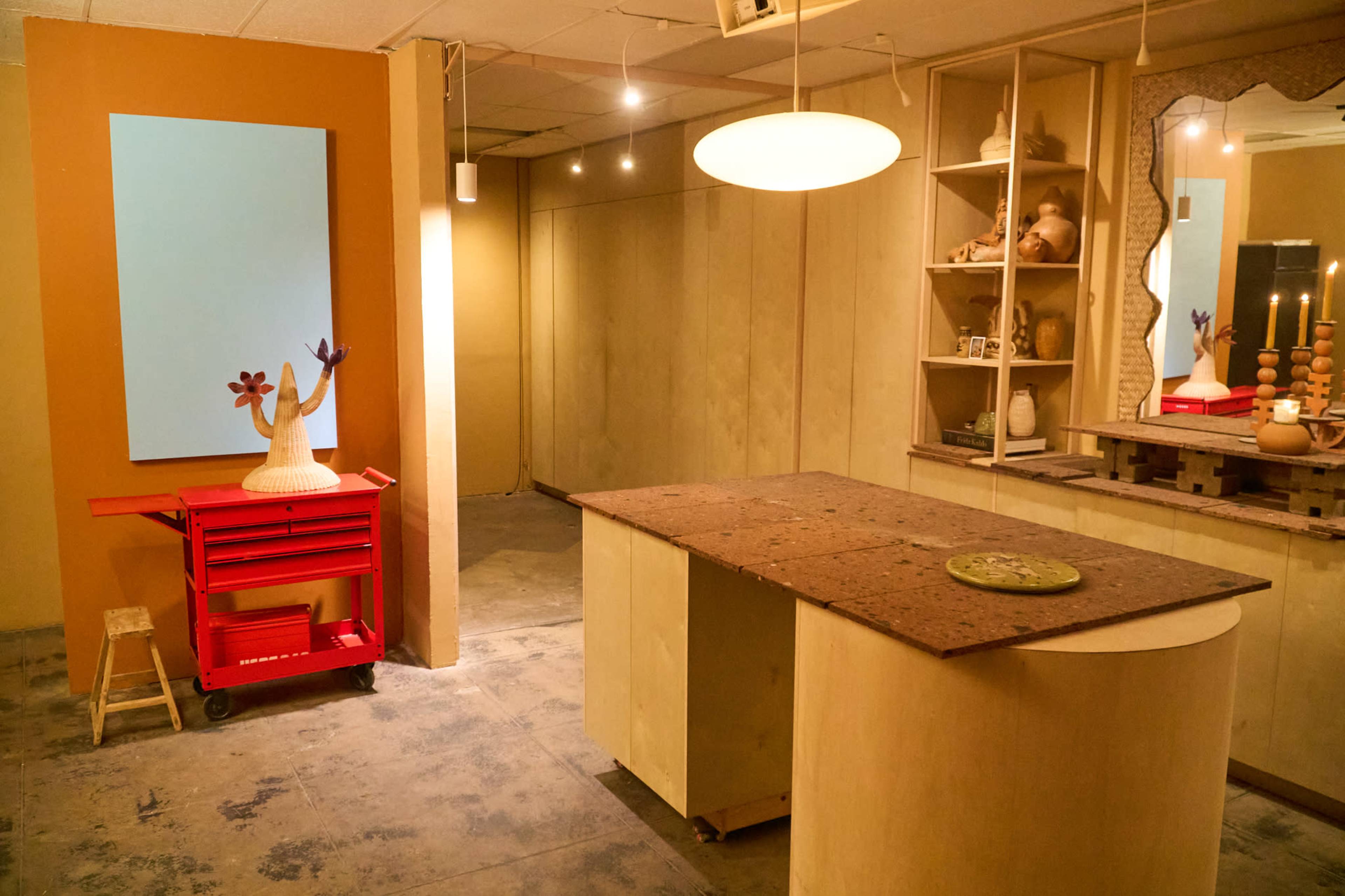 The image shows a minimalist interior space featuring an island table, a red cart, a wall mirror, and a shelf with decorative items.