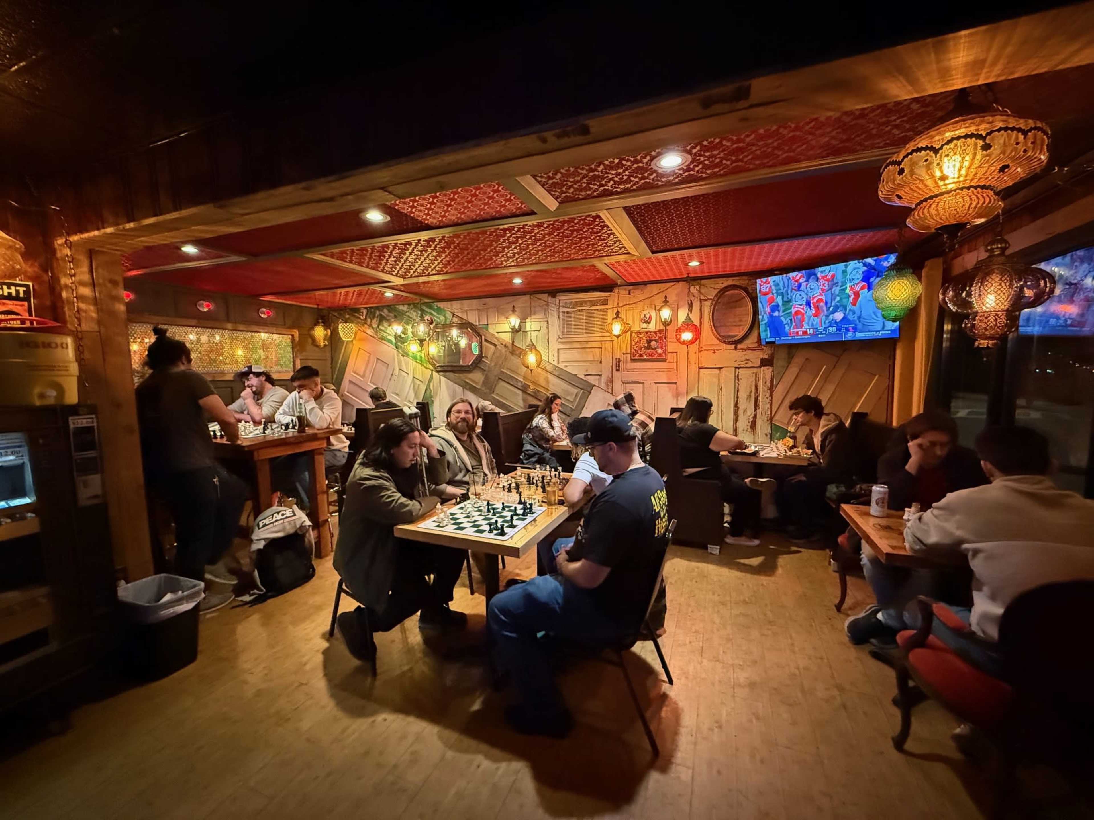 A dimly lit room with various groups of people engaged in chess games and socializing around tables decorated with colorful lights.