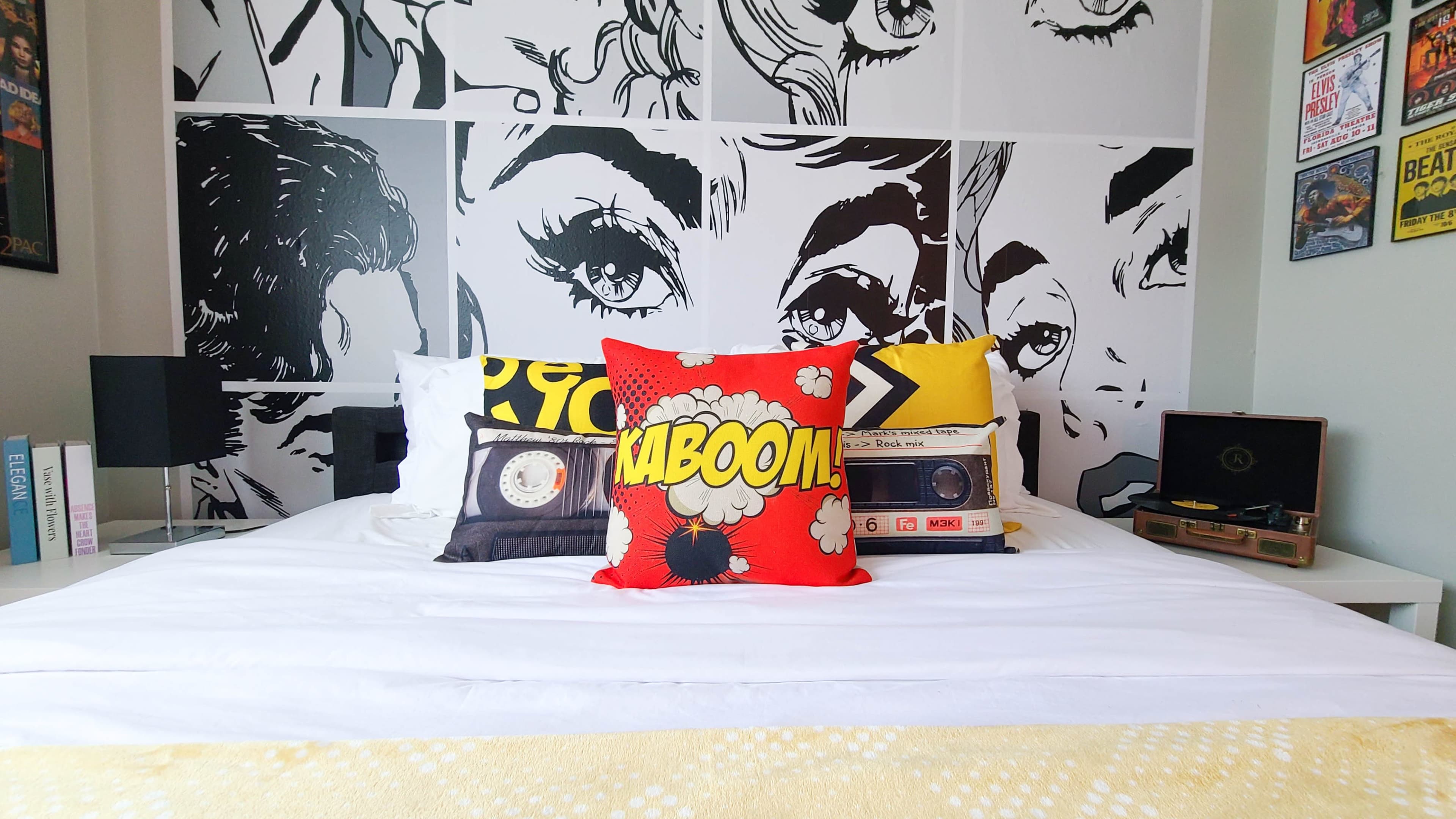 The bedroom features a bed with white linens, colorful pillows, and a backdrop of black-and-white comic-style artwork on the wall.