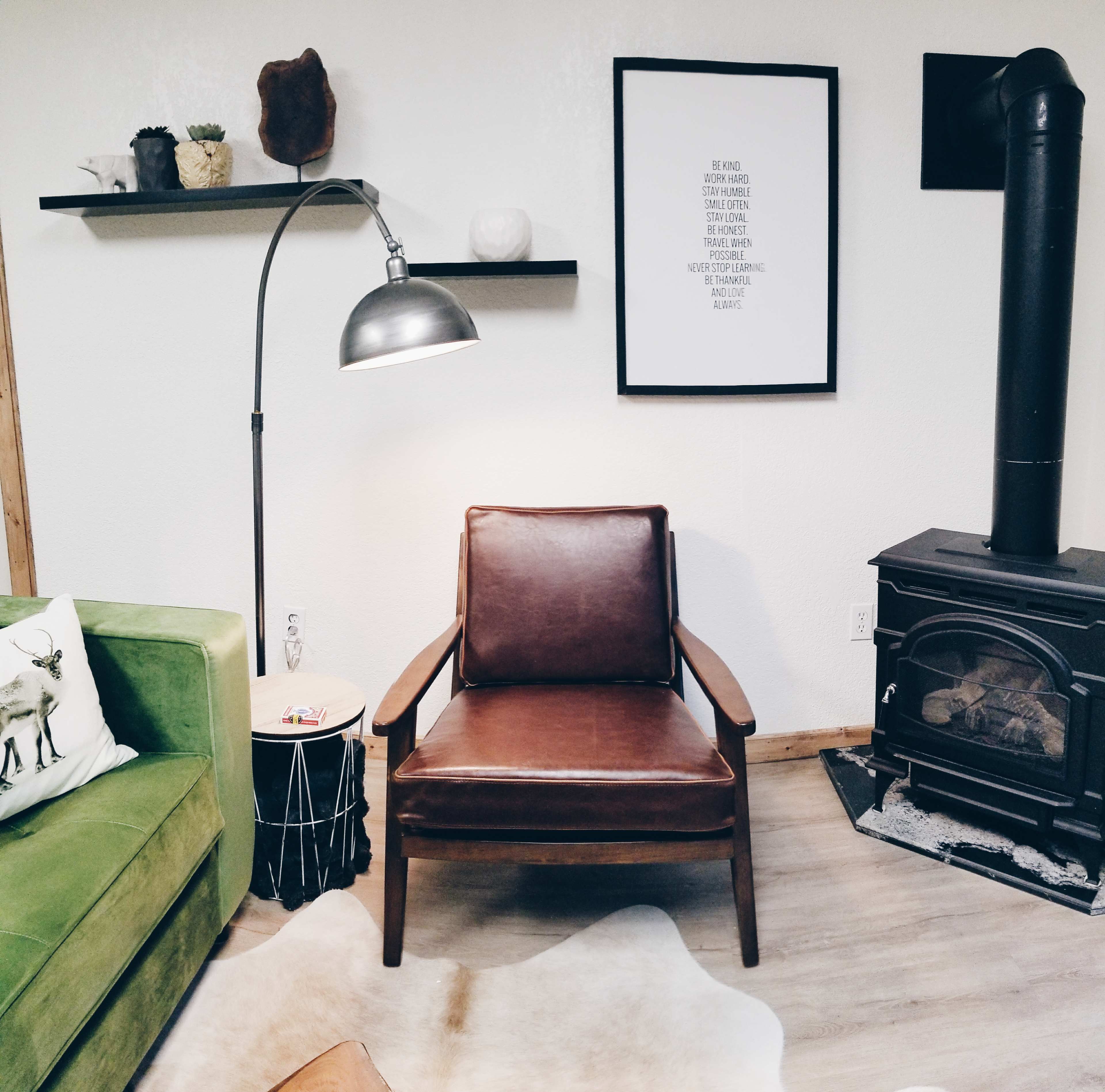 The image shows a cozy living room featuring a green sofa, a brown wooden armchair next to a floor lamp, and a wood-burning stove.