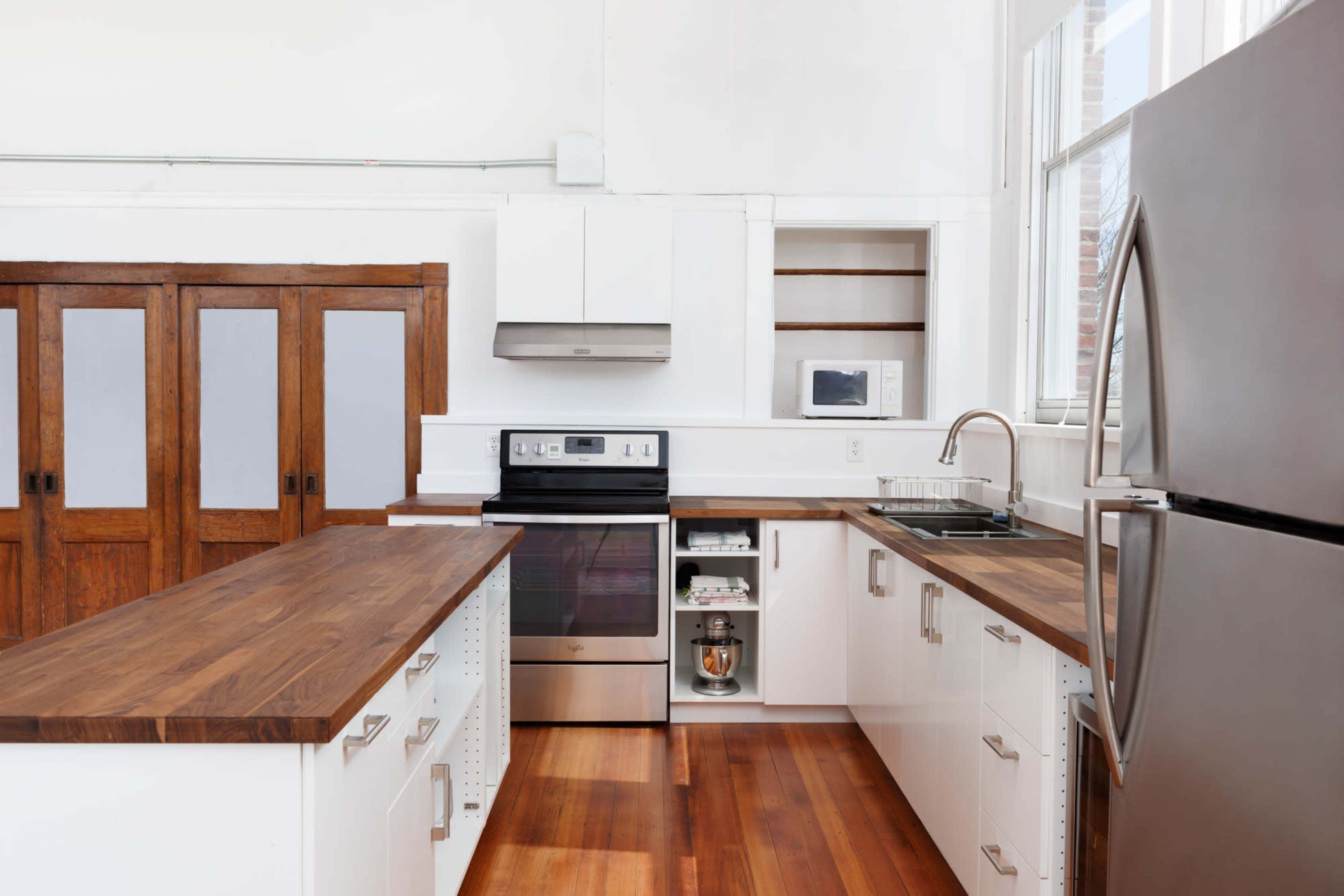 The image shows a modern kitchen featuring white cabinets, a stainless steel refrigerator, an oven, and wooden countertops, with large windows allowing natural light.