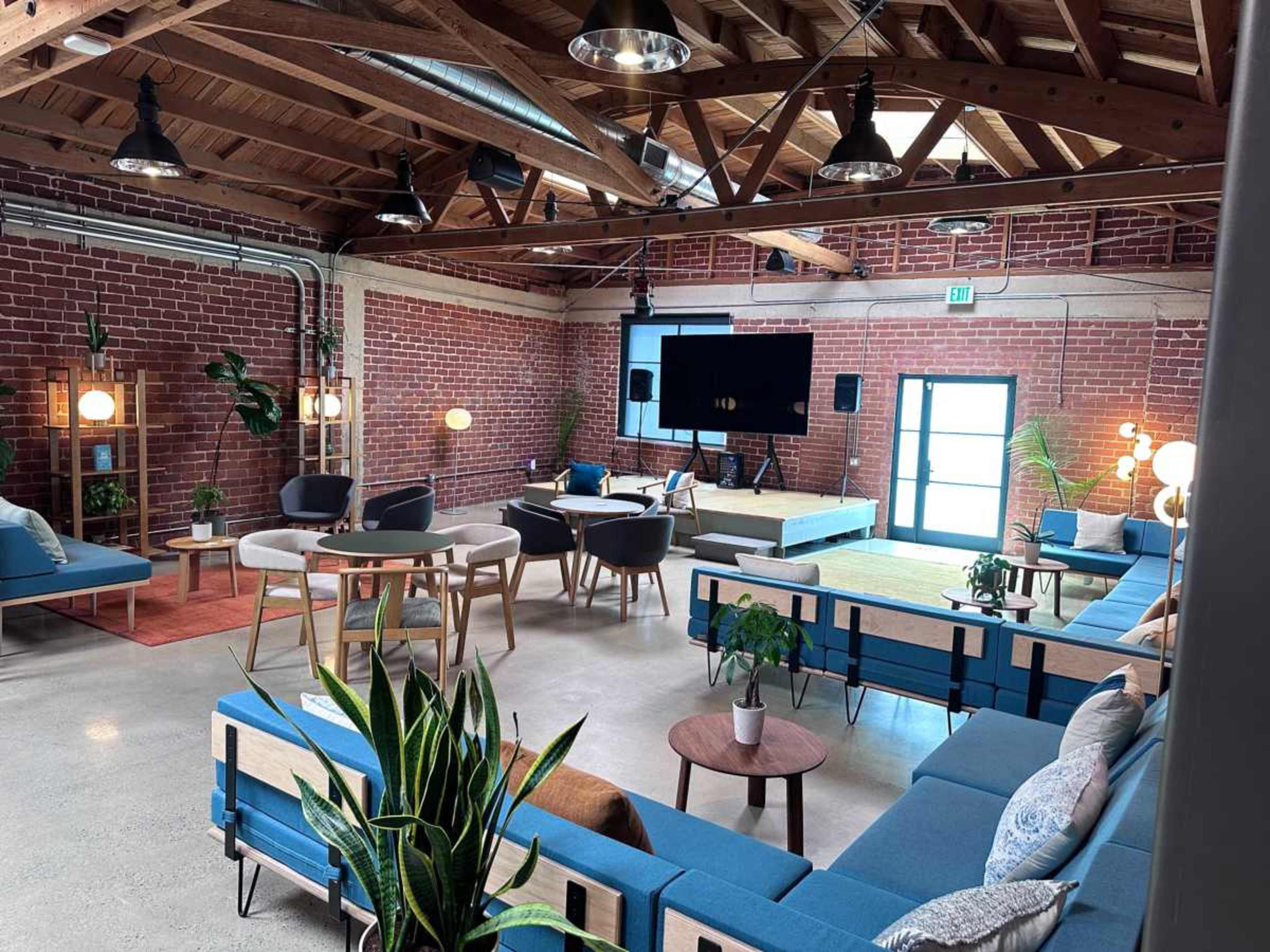 The image shows a spacious, modern loft with exposed brick walls, wooden beams, and various seating arrangements including couches, chairs, and tables.