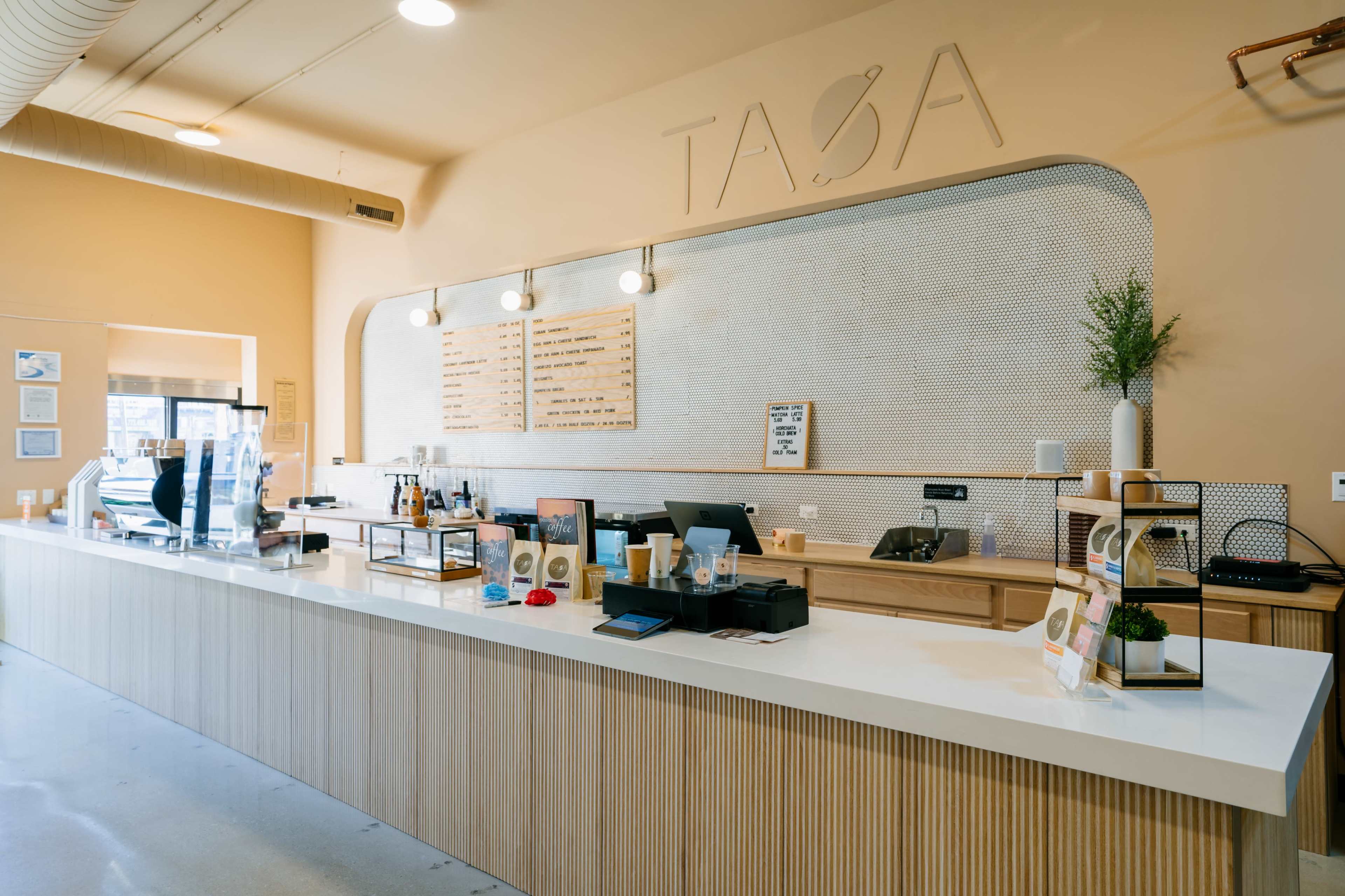 The image depicts a minimalist café counter with a light wood finish, a cash register, and various menu items displayed on the wall behind it.