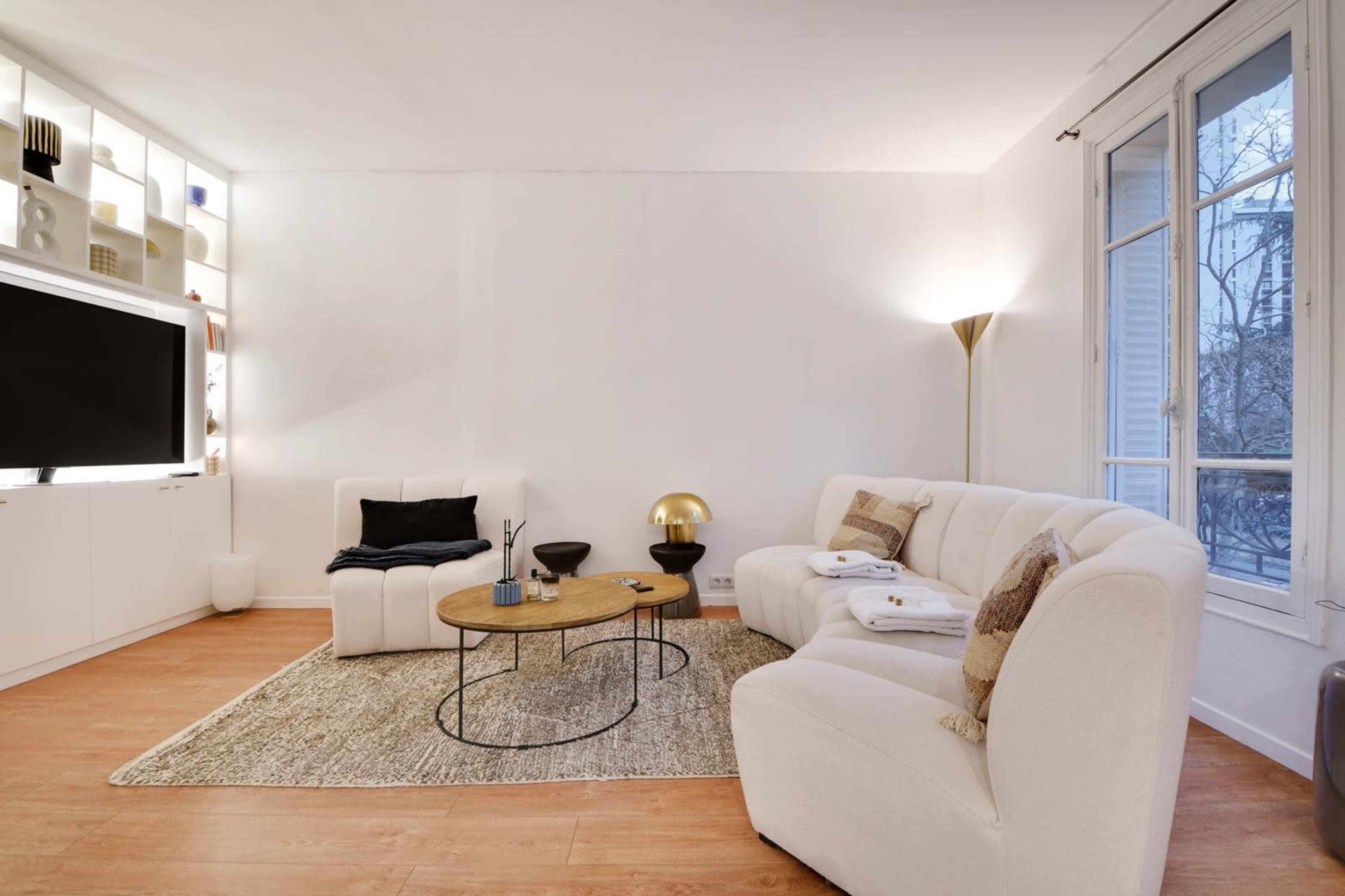 A modern living room features a white sectional sofa, a round wooden coffee table, and a floor lamp, with a large TV mounted on the wall and a window allowing natural light.