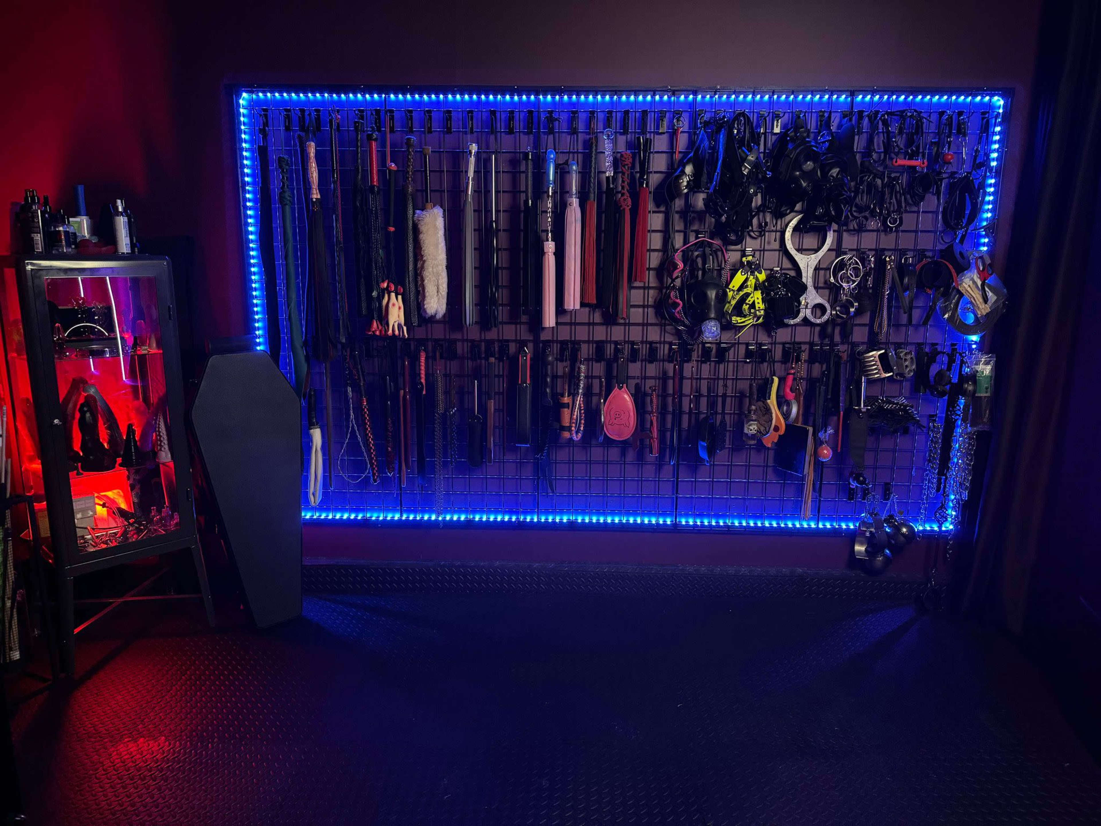 The image shows a wall-mounted rack with various tools and equipment, illuminated by neon blue lights, alongside a display case containing additional items.