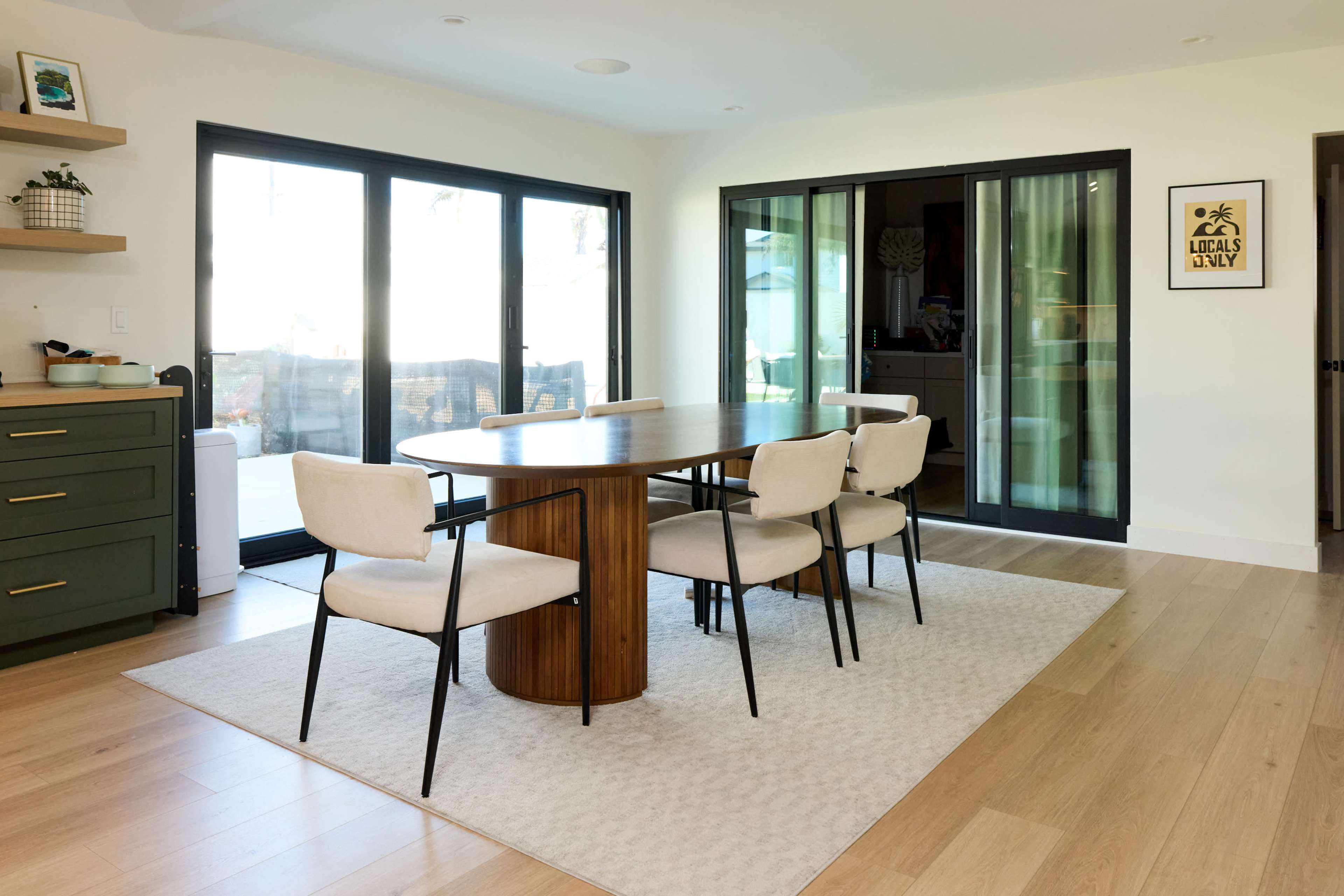 A modern dining area features a round wooden table surrounded by four beige chairs, with large glass doors providing natural light and access to an outdoor space.