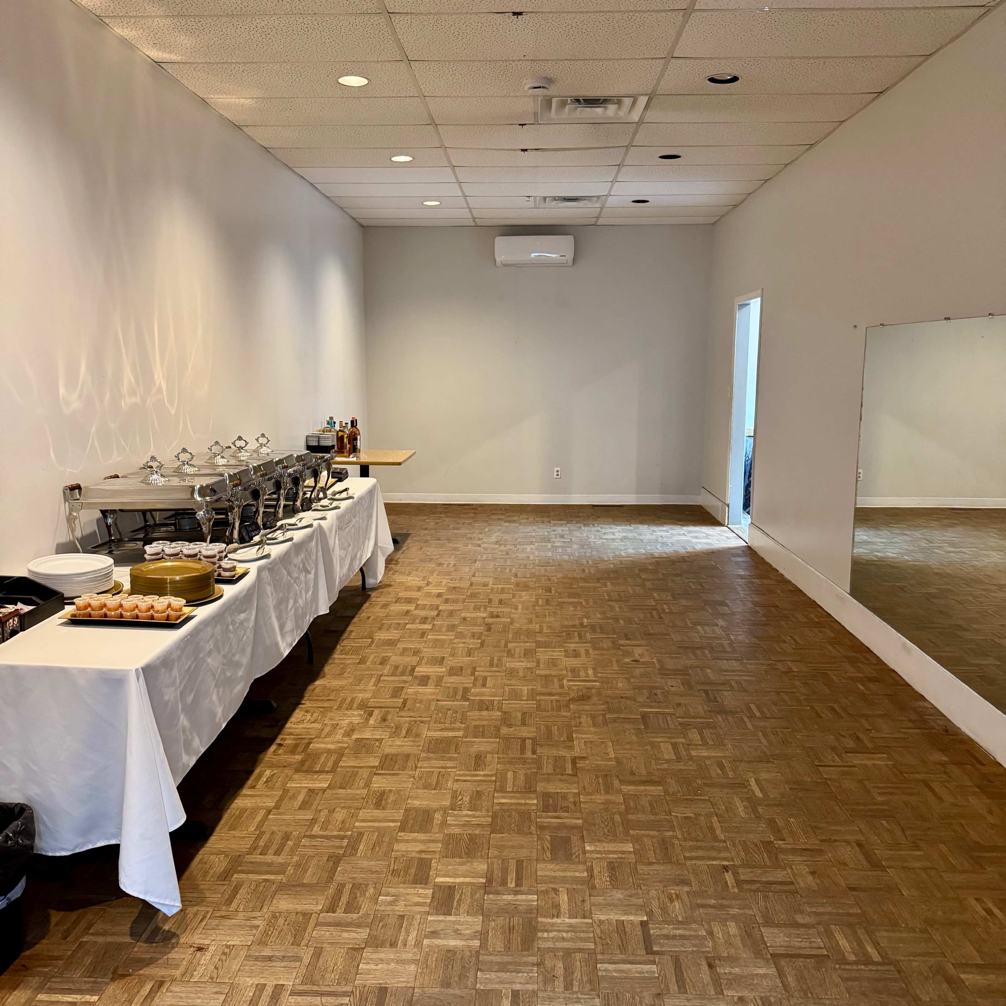 The image shows a spacious room with a wooden floor, featuring a table laid out with food and drinks on one side and a mirror on the opposite wall.