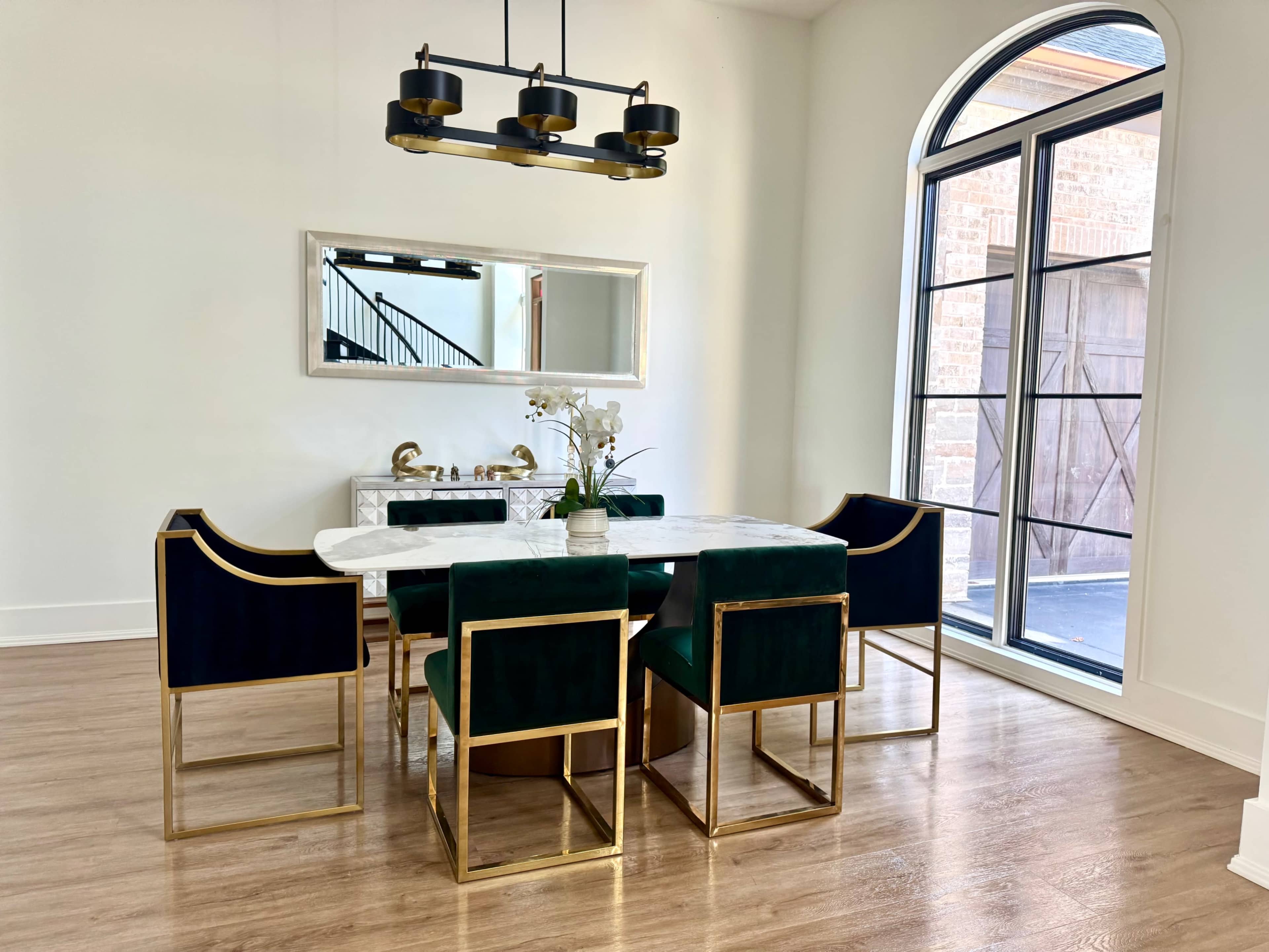 The image shows a modern dining room featuring a marble-topped table surrounded by six green and gold chairs, with large windows letting in natural light.