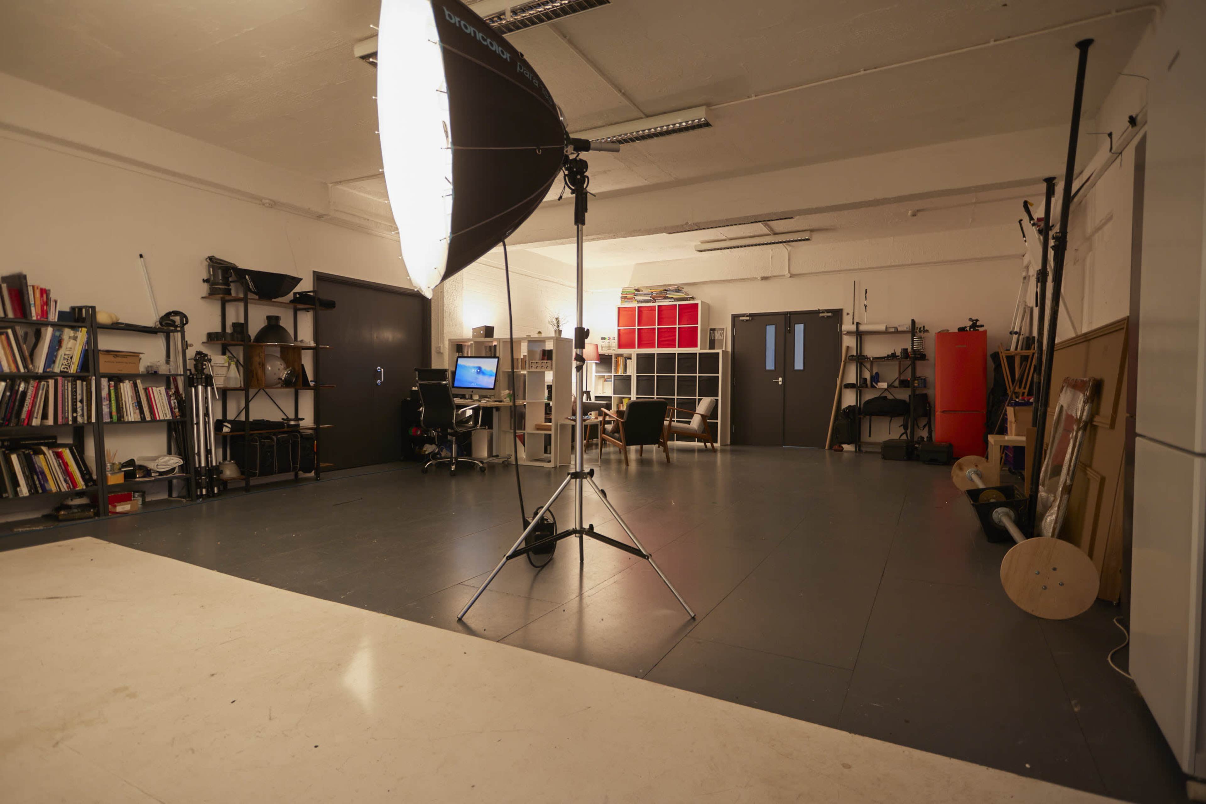 A large, empty studio space equipped with a photography light, shelves filled with books, a desk with a computer, and fitness equipment against the walls.