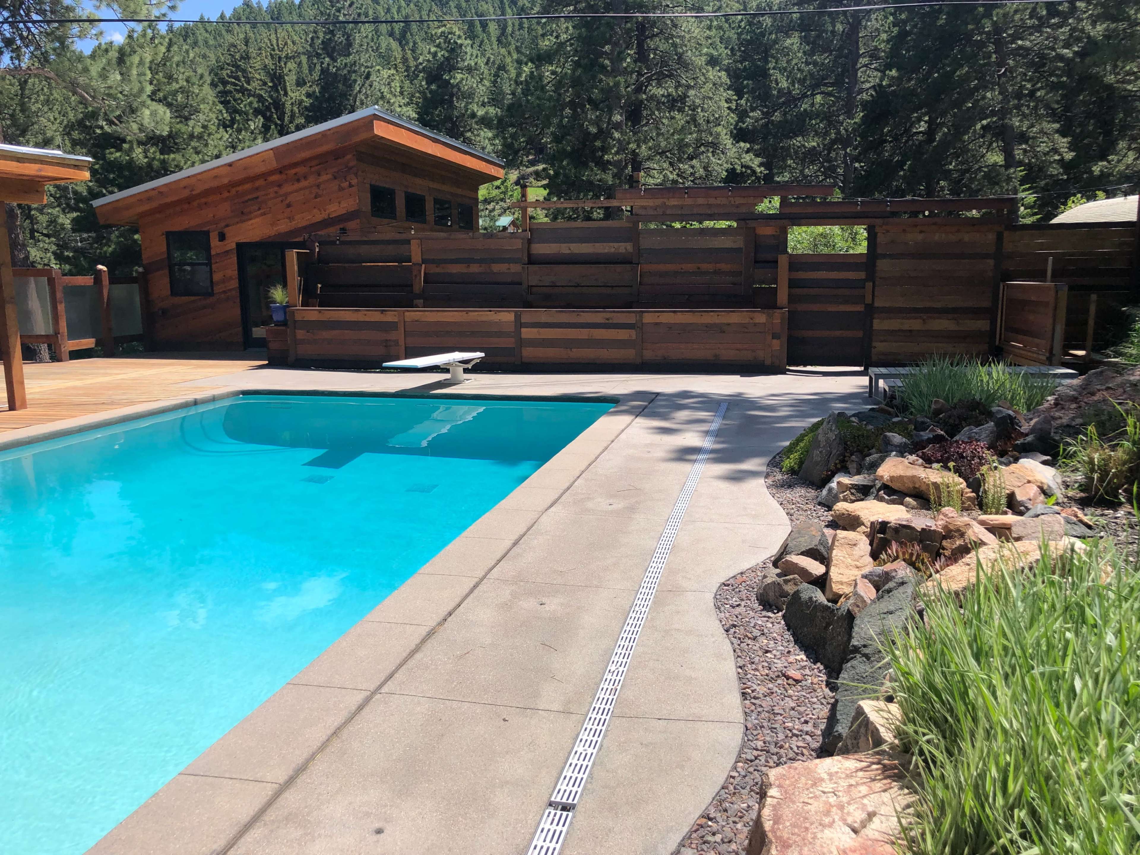 The image shows a rectangular swimming pool beside a wooden deck and modern cabin set against a backdrop of trees.