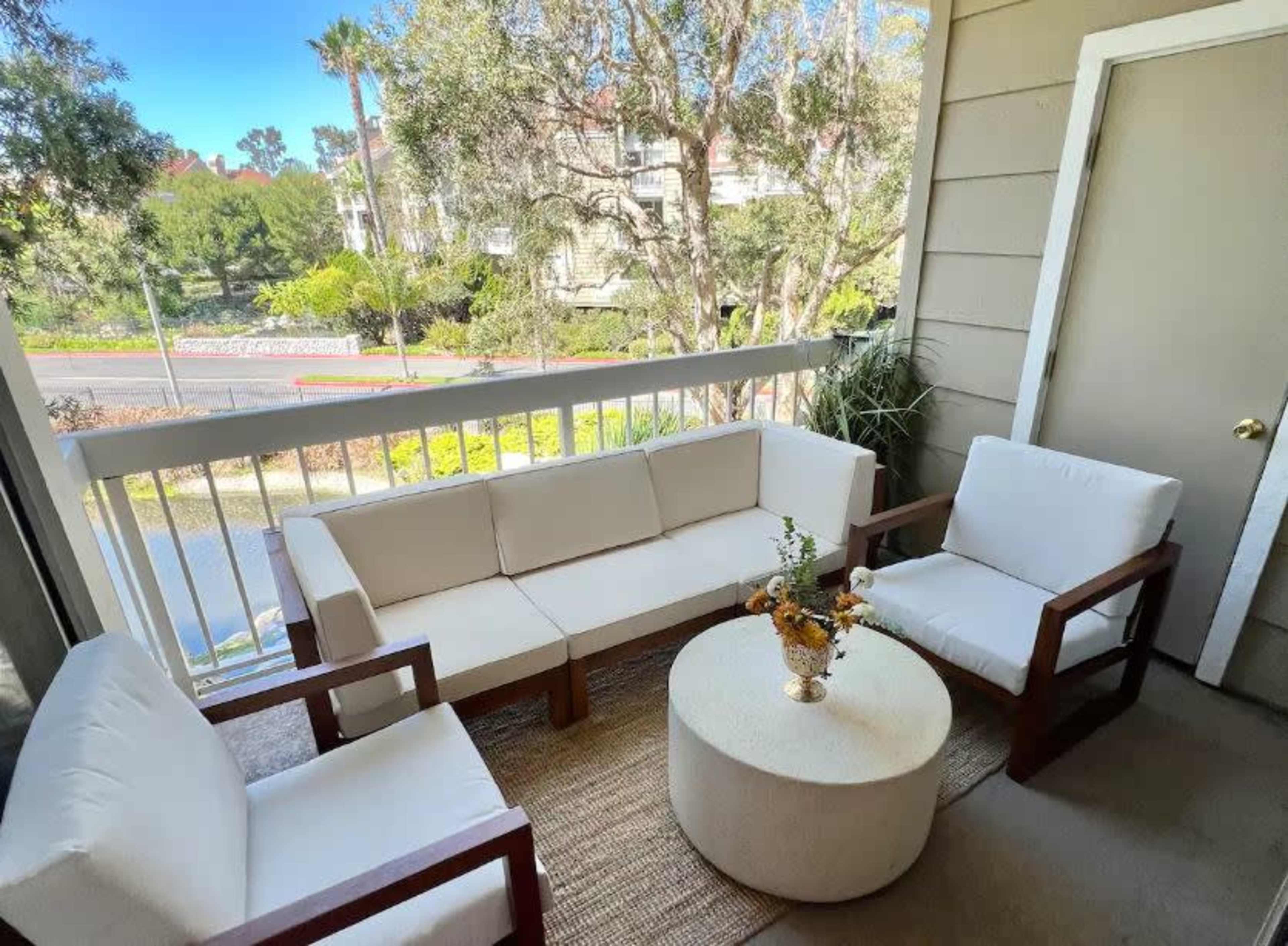 A cozy balcony seating area with a white outdoor sofa and two chairs arranged around a circular coffee table, surrounded by greenery.
