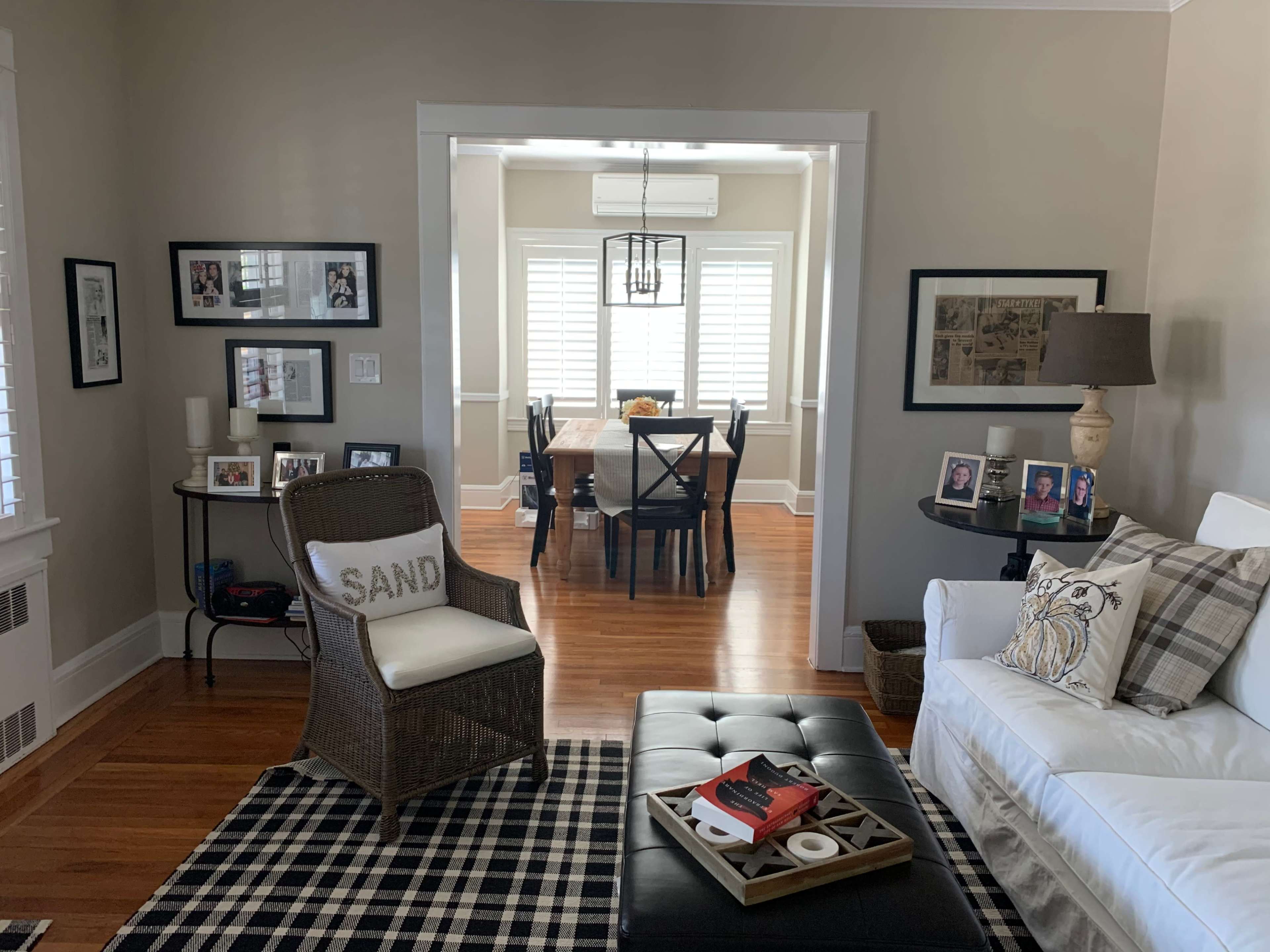 A living room features a white sofa, a black ottoman on a checkered rug, and a view into a dining area with a wooden table and window shutters.