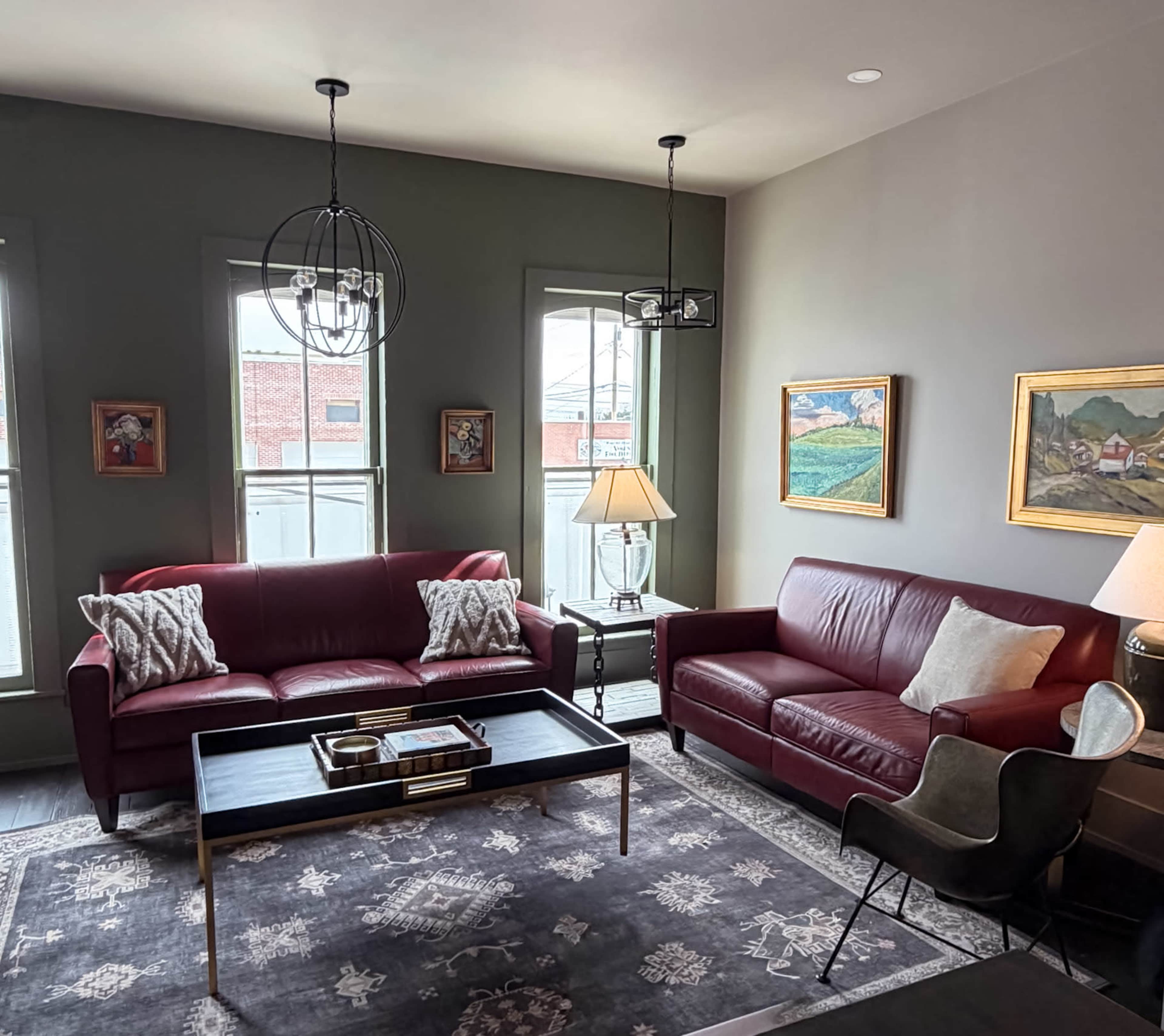 The image shows a modern living room with two red leather sofas, a coffee table, an area rug, and wall art, illuminated by natural light from large windows.