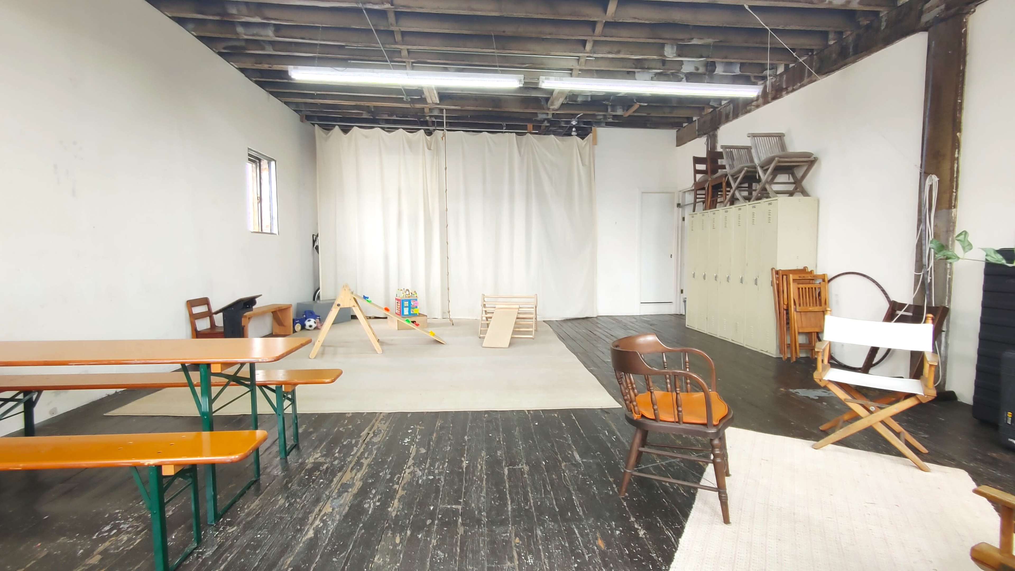 The image shows a spacious room with wooden floors, a few tables and chairs arranged, and equipment for play set up in the center, along with metal lockers against one wall.
