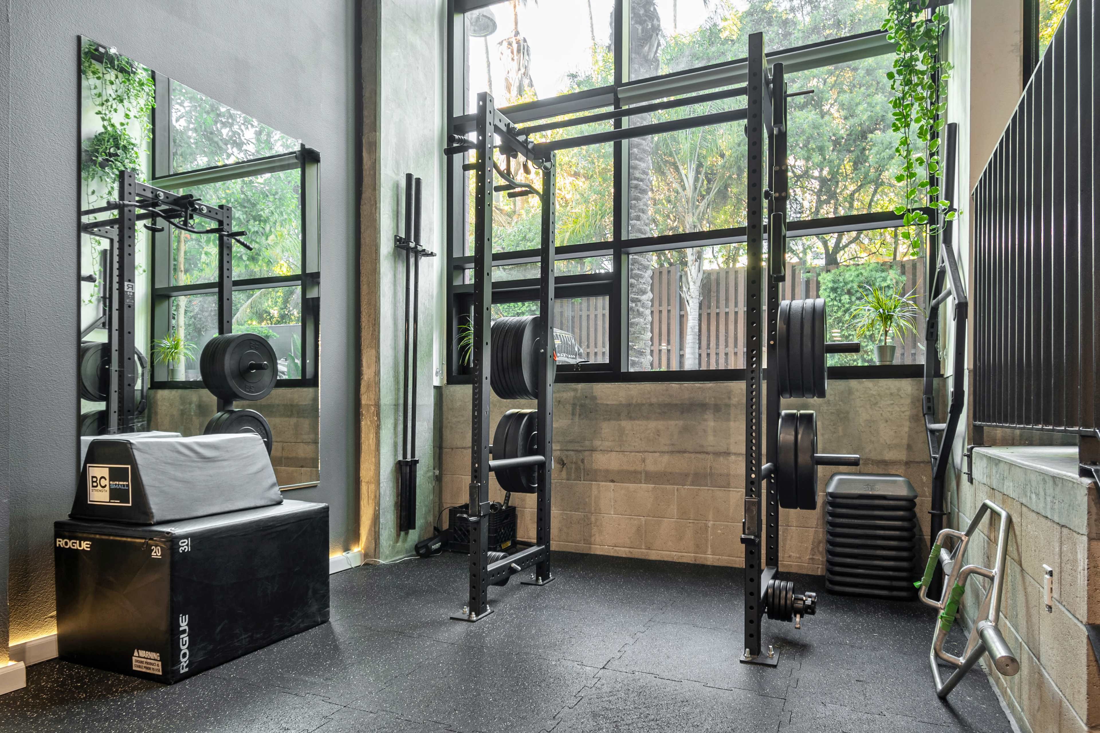 A modern gym area featuring a weightlifting rack, a black exercise box, and various weight plates in a bright space with large windows.