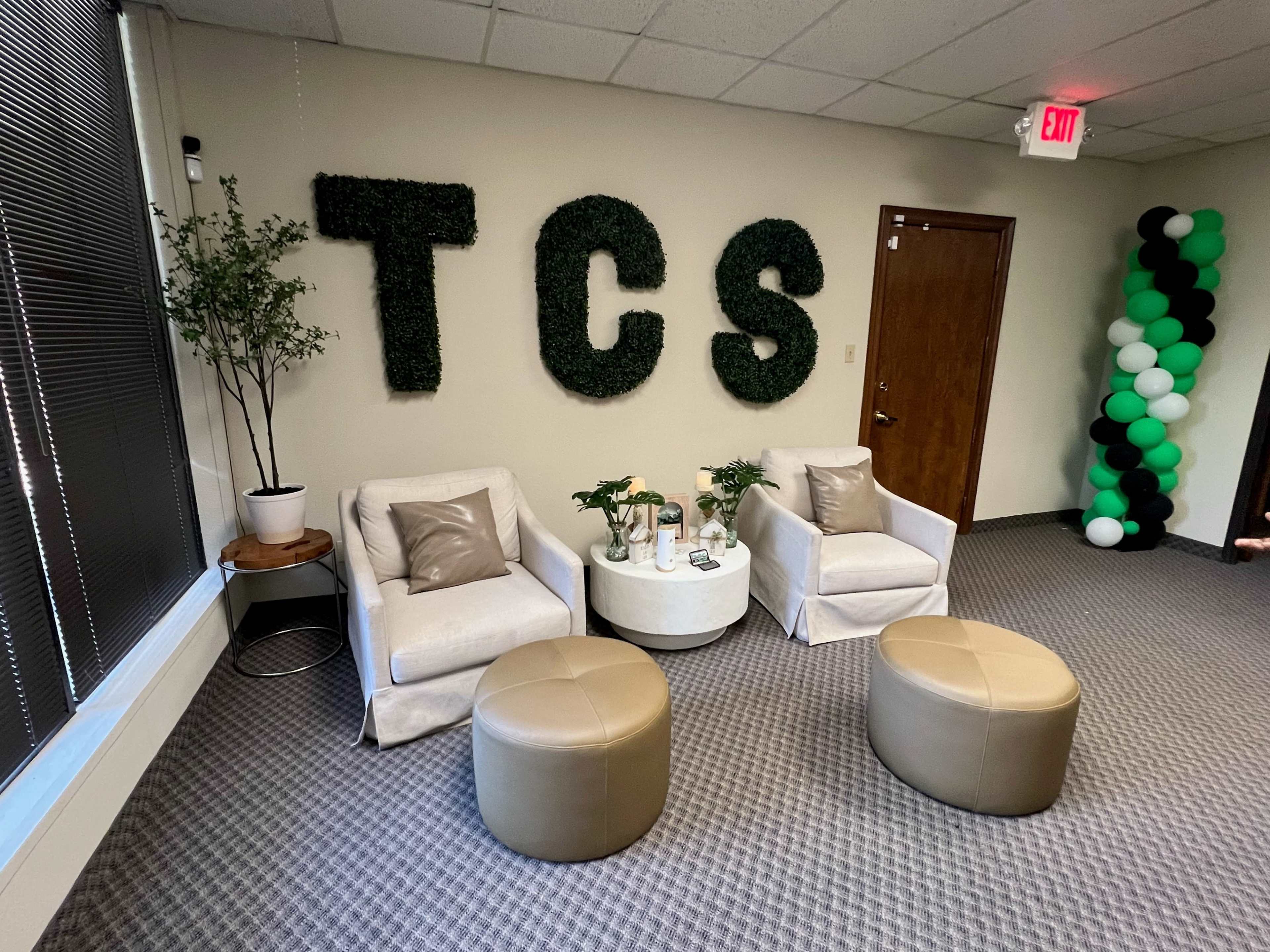 The image shows a cozy lounge area with two armchairs, a round coffee table, and decorative plants, featuring the letters "TCS" made from greenery on the wall.