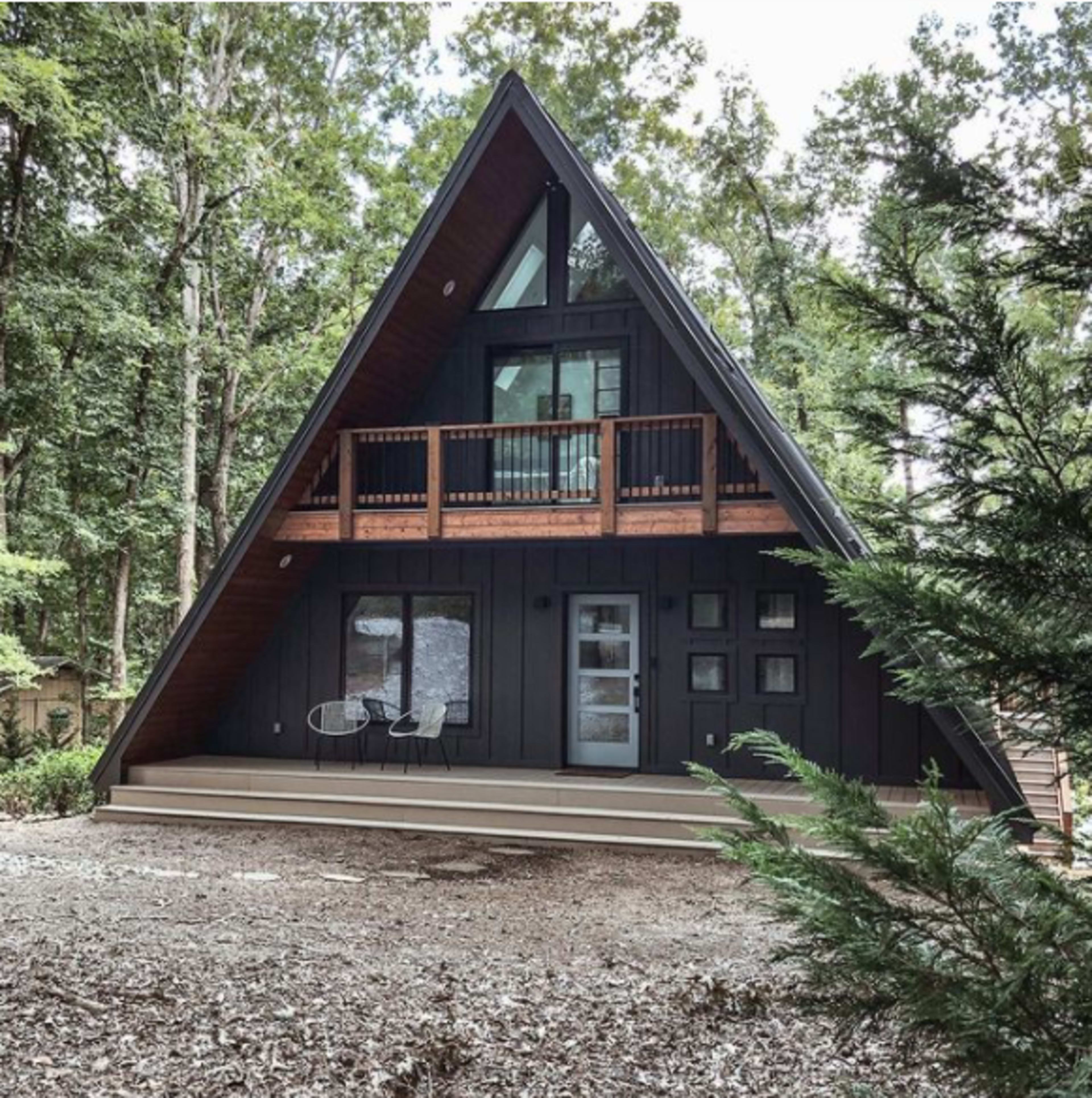 A modern A-frame house surrounded by trees, featuring a porch with seating and large windows.