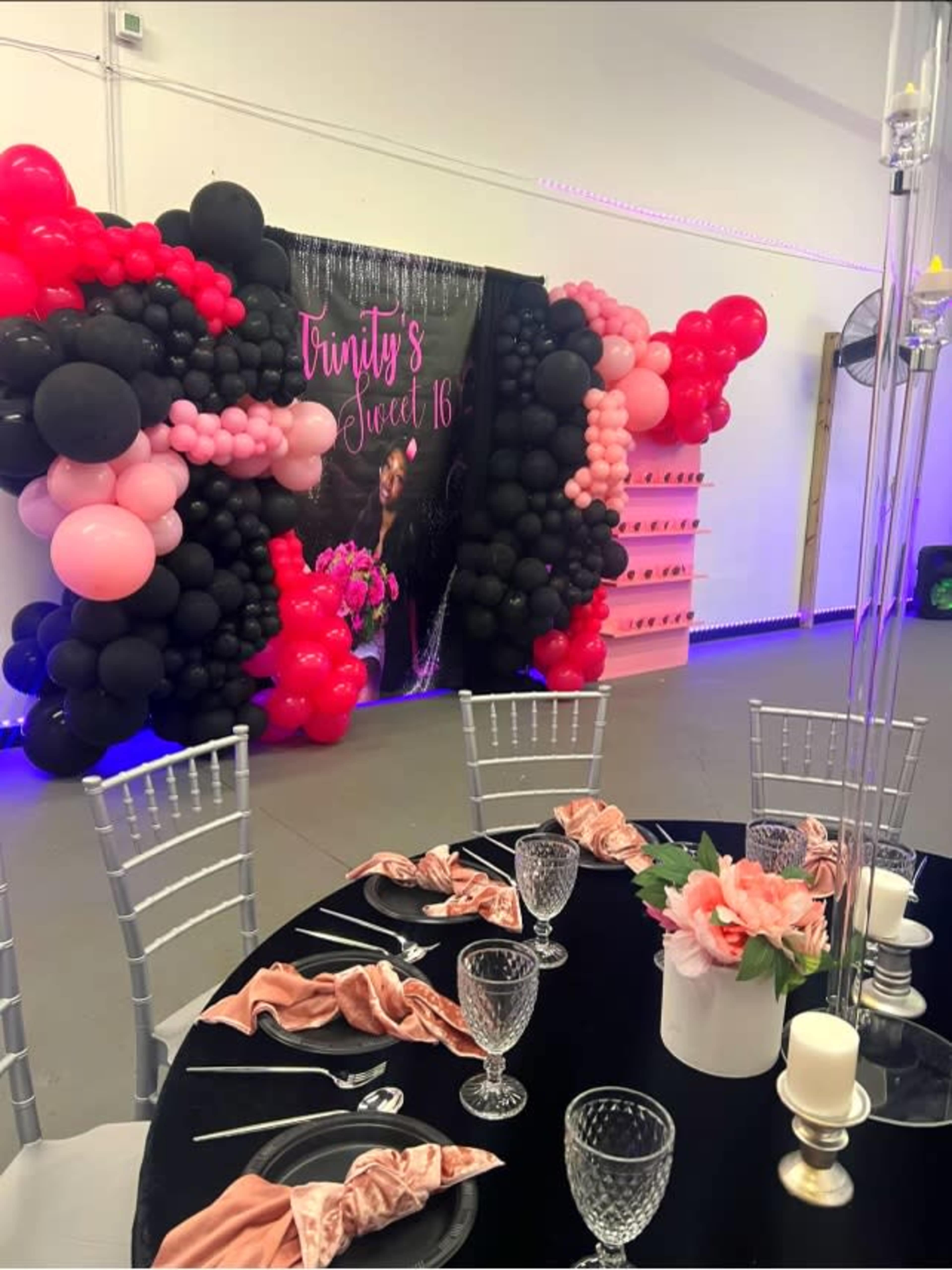 The image shows a decorated event space for a Sweet 16 celebration, featuring a wall of black, pink, and red balloons, a large banner, and a table set with plates, glasses, and napkins.
