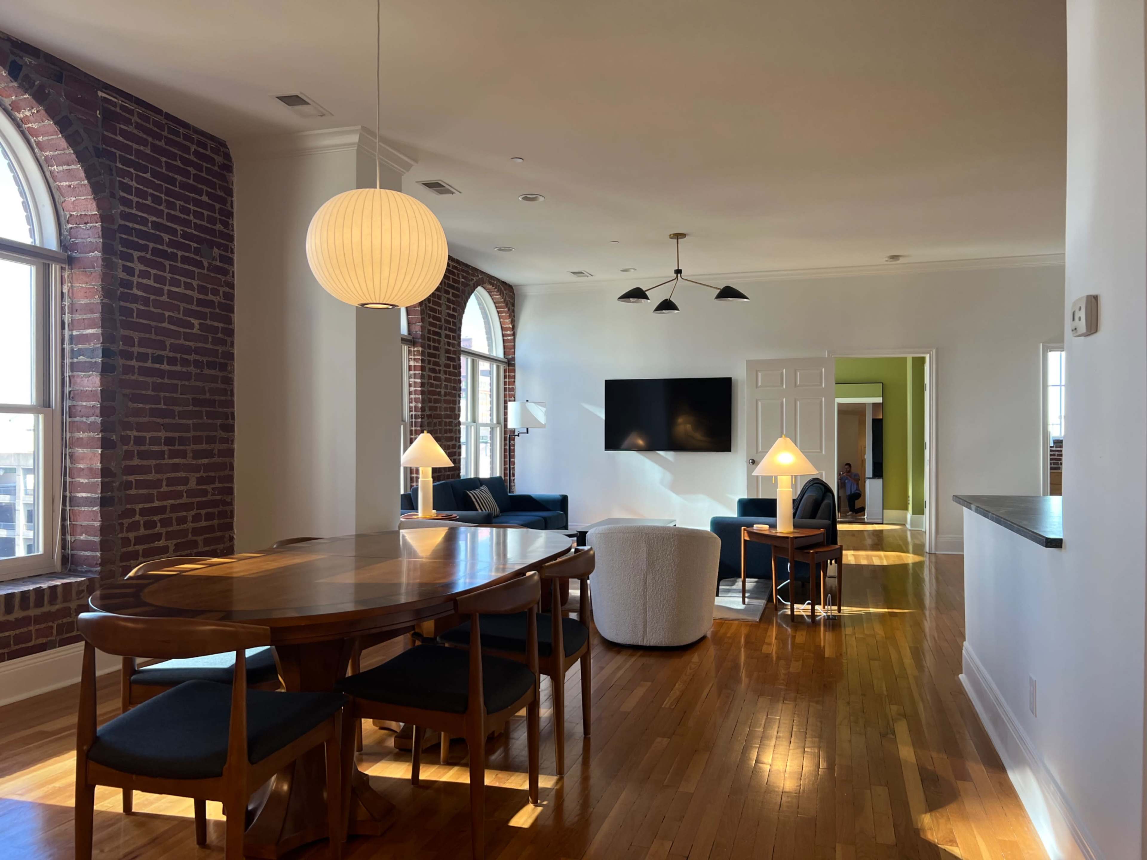 The image shows a spacious living area with a dining table, a seating area featuring a sofa and chairs, and large windows allowing natural light to illuminate the room.