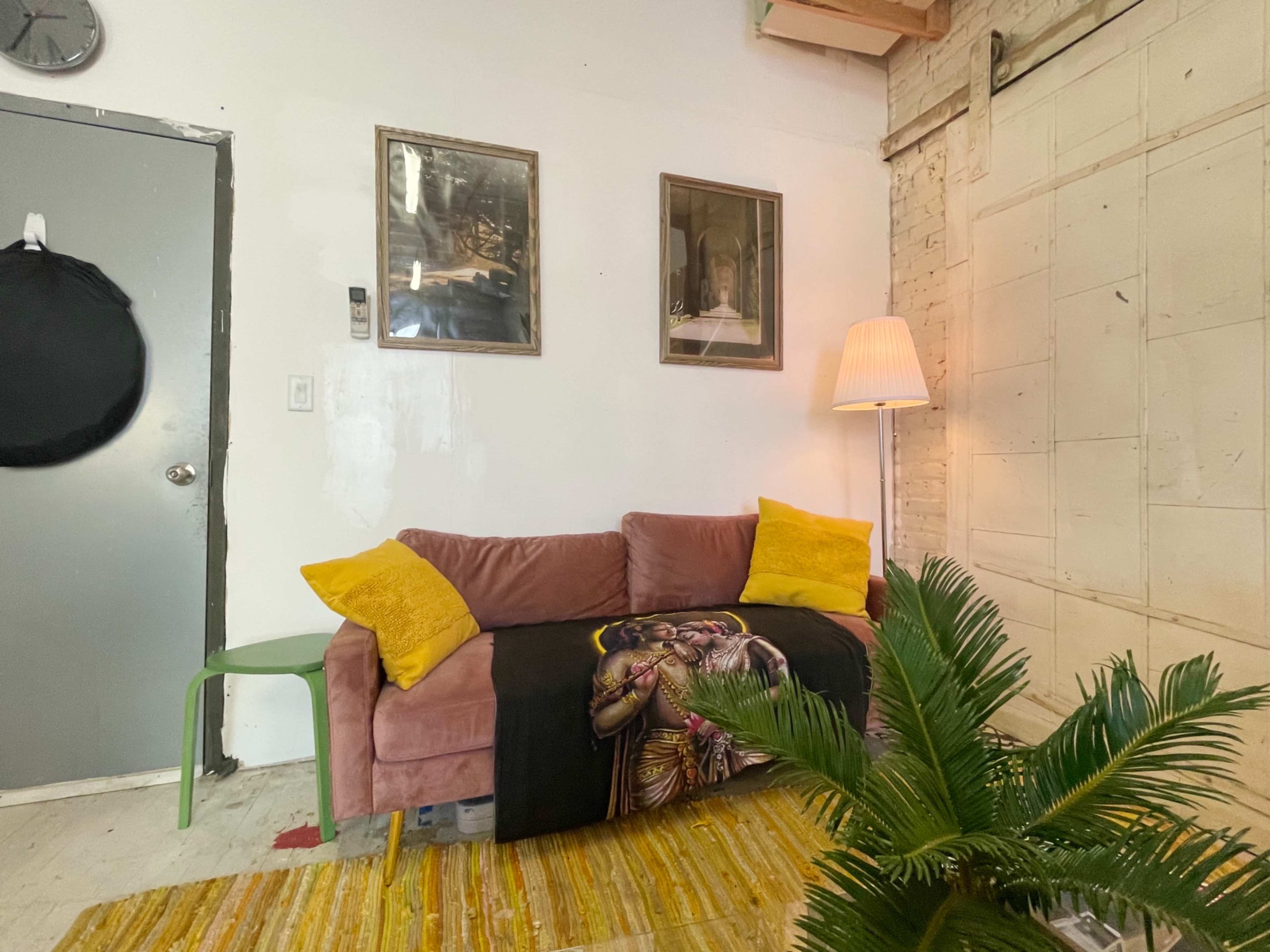 The image shows a small lounge area with a pink sofa, two framed pictures on the wall, a lamp beside the sofa, and a green chair in front of a potted plant.
