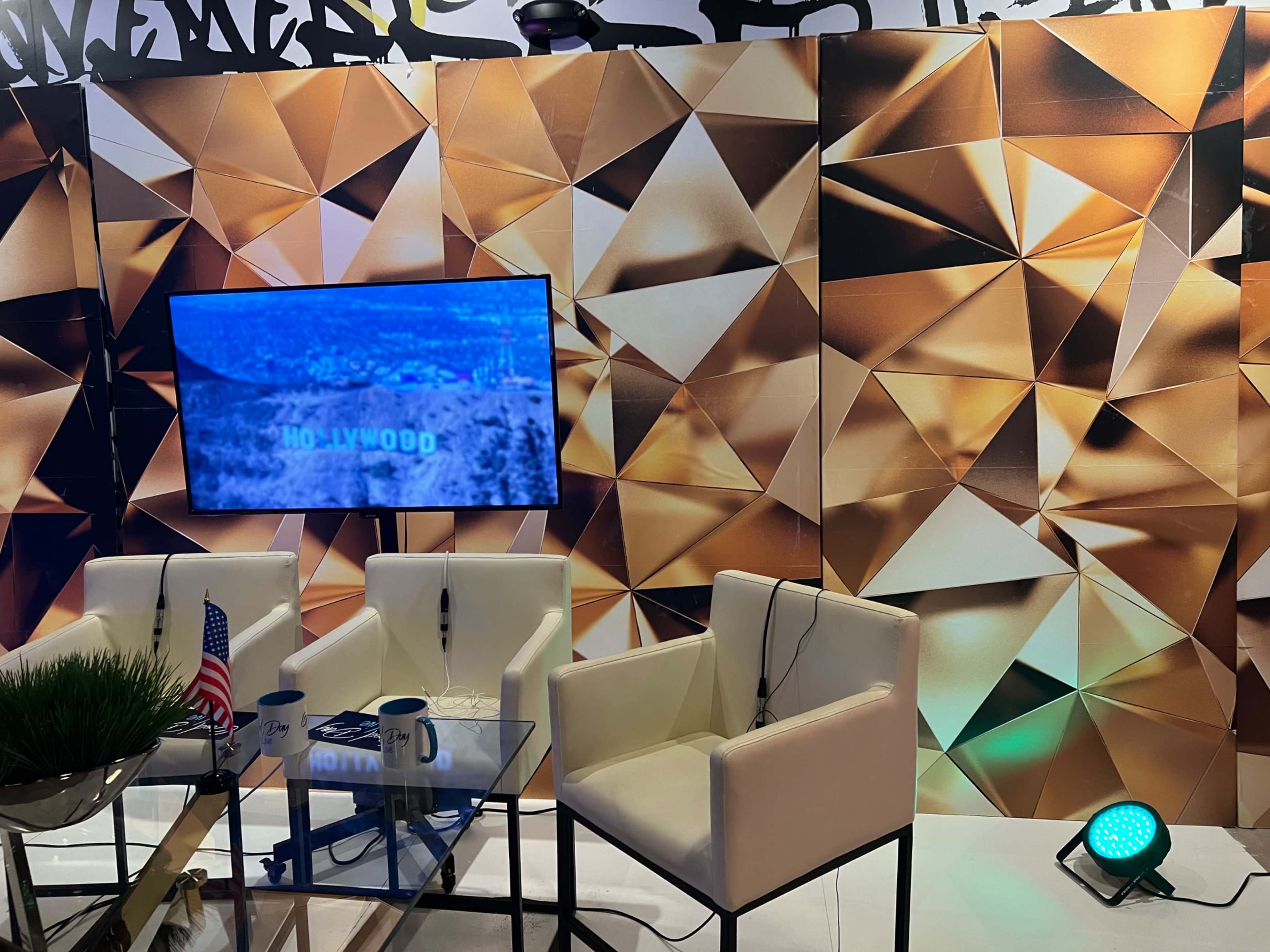 A modern set design featuring a television displaying the word "HOLLYWOOD," surrounded by a geometric gold-patterned backdrop, with white chairs and a table setup.